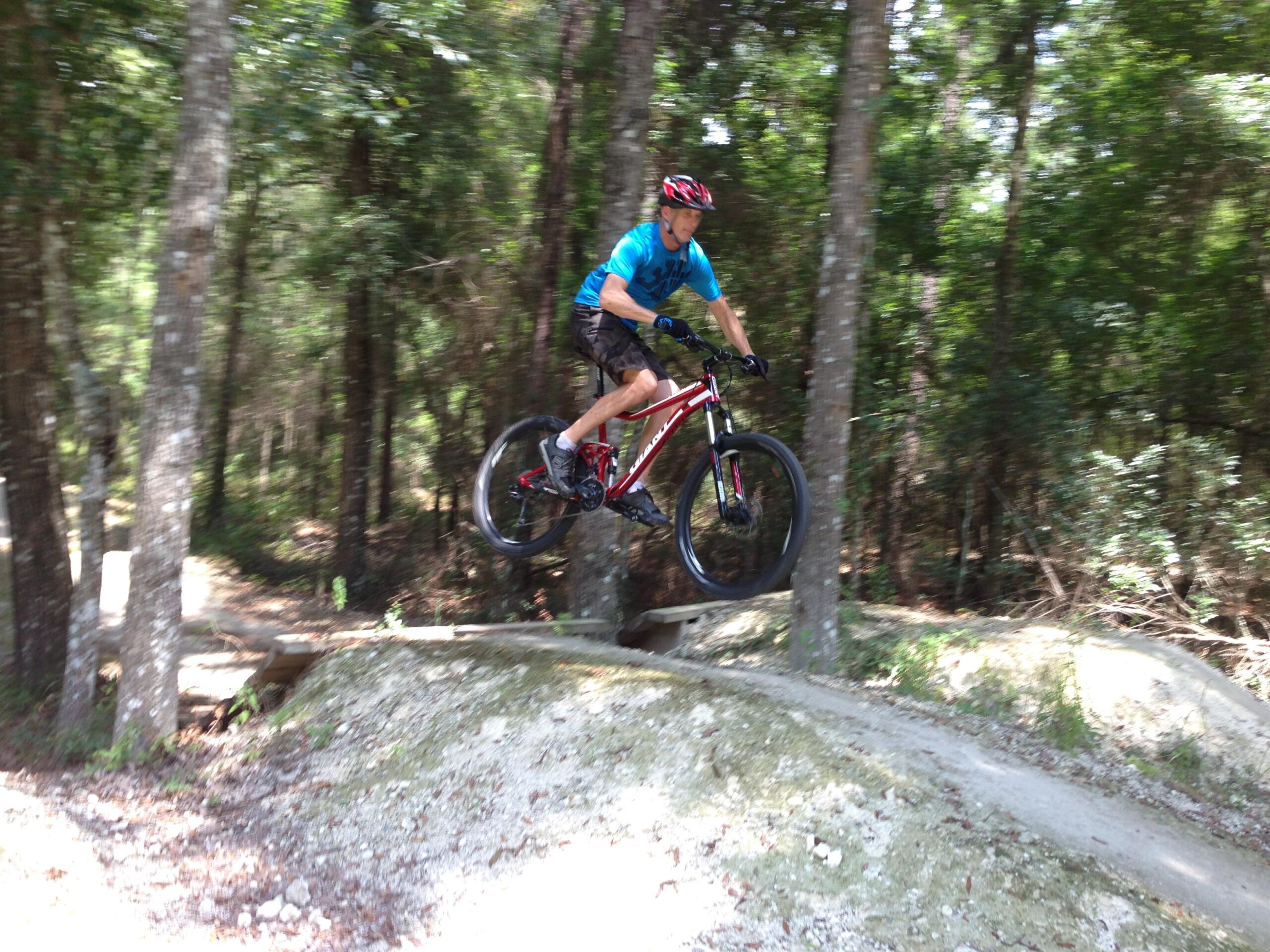 Giant Trance: A young boy riding a mountain bike while jumping off a dirt ramp in a wooded area, surrounded by tall trees and greenery. The photo captures a sense of motion and excitement as the bike is airborne.