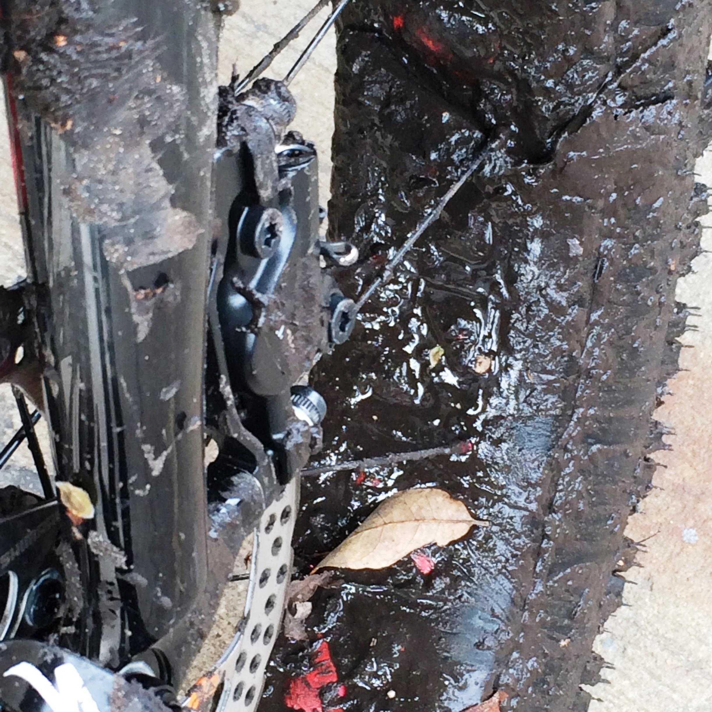Close-up view of a mountain bike tire and rear brake system covered in mud, with leaves scattered on the ground. The image captures the bike's dirty components, highlighting a rugged terrain experience. Environmental Center mountain bike trail.