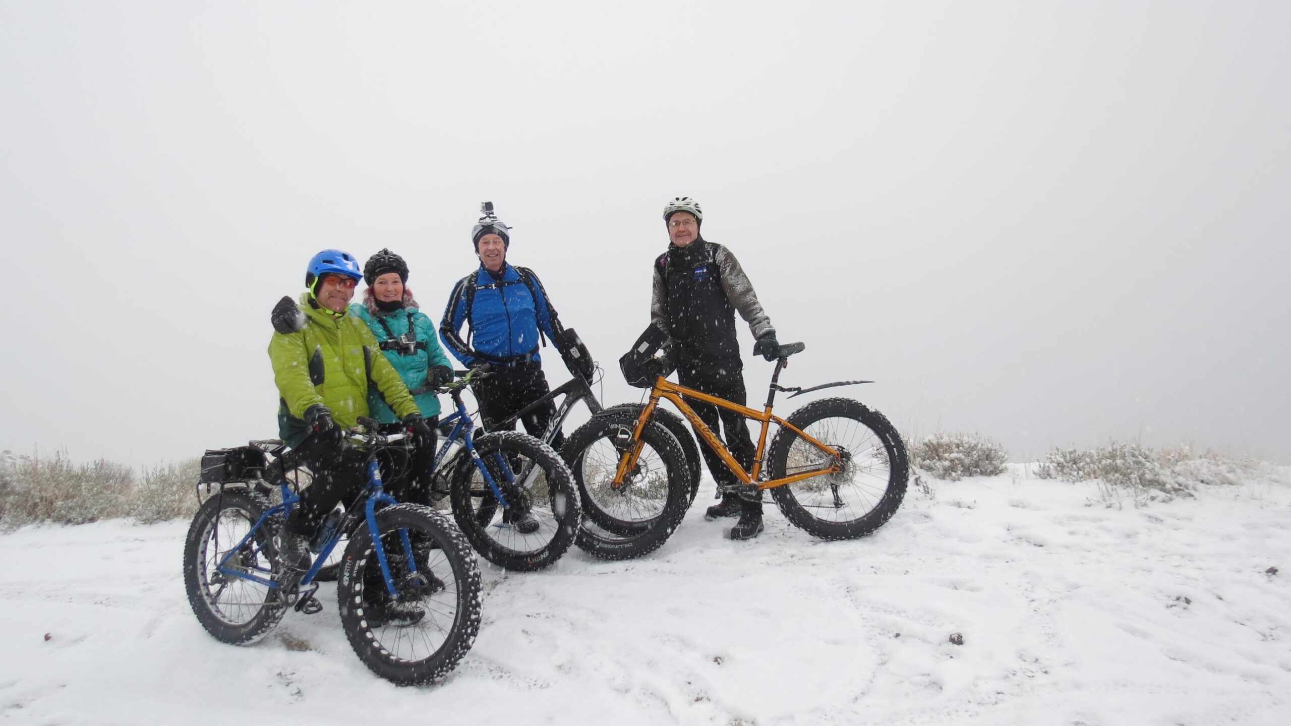 Four cyclists pose with their bikes in a snowy, foggy landscape. They are dressed in winter cycling gear, with two riding fat bikes and two on traditional mountain bikes. The background is obscured by mist and falling snow, emphasizing the chilly outdoor conditions of their ride. Fire Road down to Kamloops Lake mountain bike trail.