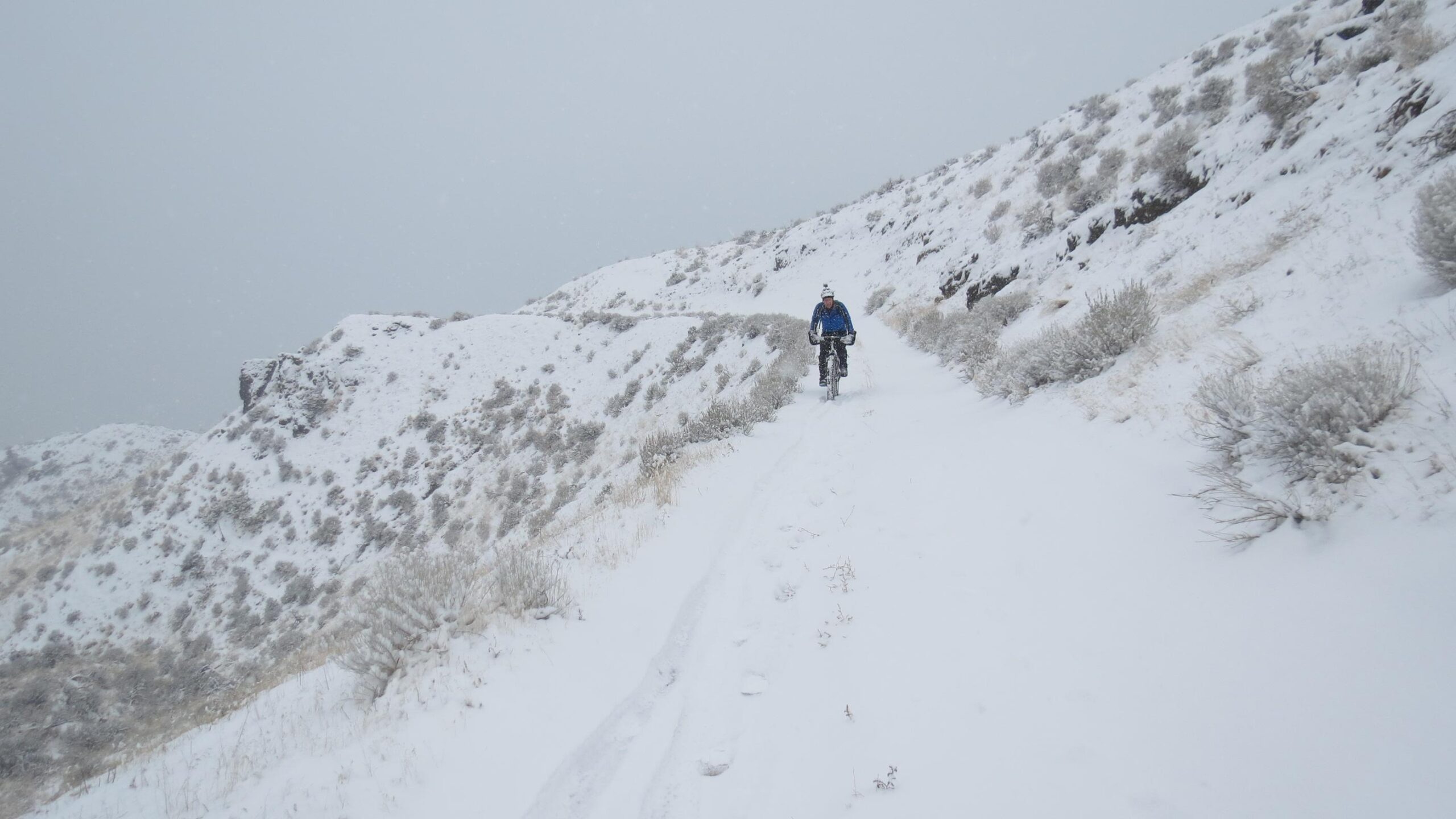 A cyclist rides along a snowy trail in a mountainous landscape, surrounded by gently sloping hills covered in a thick layer of snow. Light snowfall adds a serene atmosphere to the scene, with patches of grass and shrubs peeking through the white covering. The sky is overcast, creating a soft, diffused light. Fire Road down to Kamloops Lake mountain bike trail.