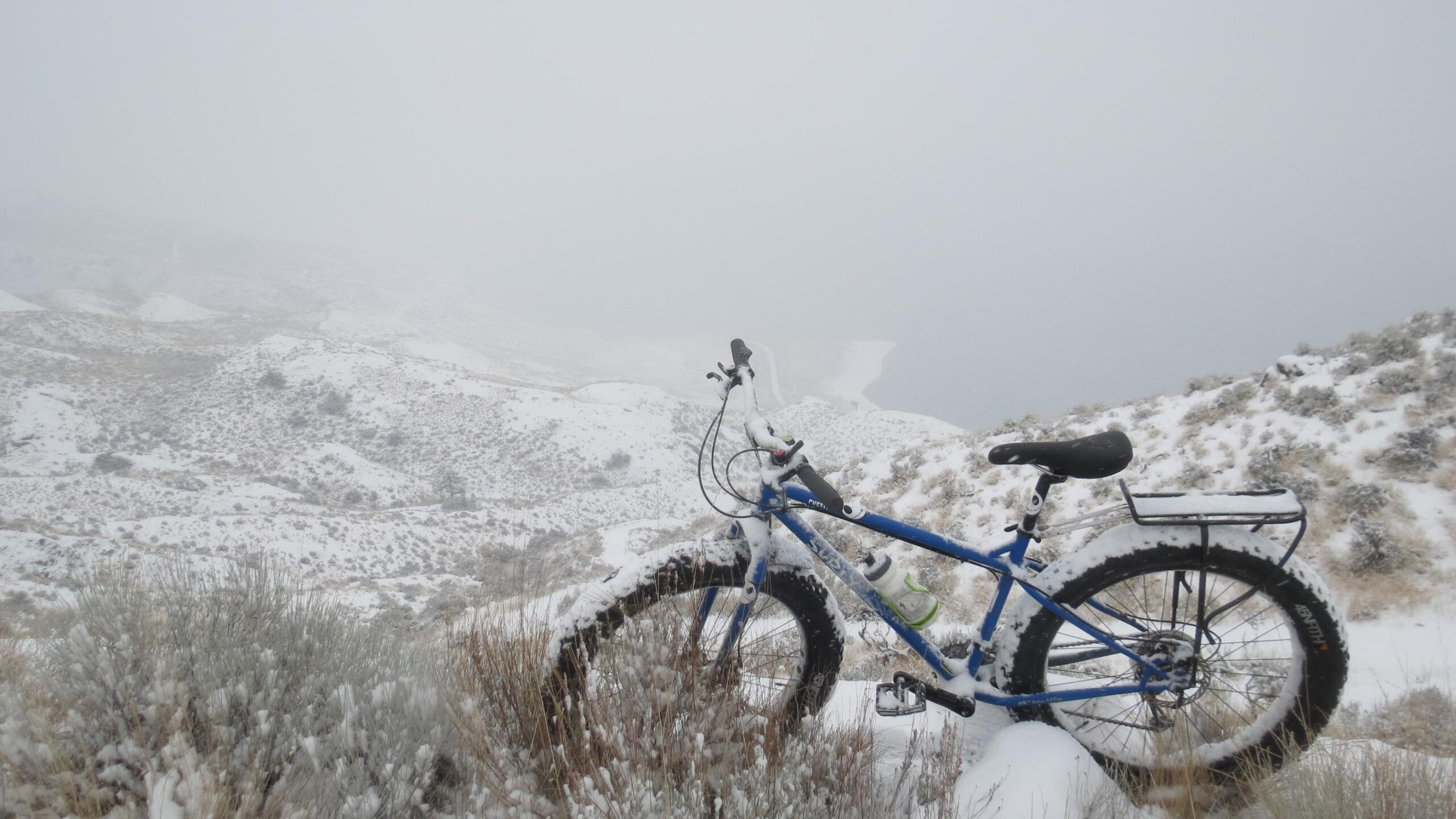 A blue fat bike covered in snow is parked on a snowy hillside, surrounded by a winter landscape featuring rolling mounds and sparse vegetation. The scene is shrouded in fog, creating a misty atmosphere with limited visibility in the background. Fire Road down to Kamloops Lake mountain bike trail.