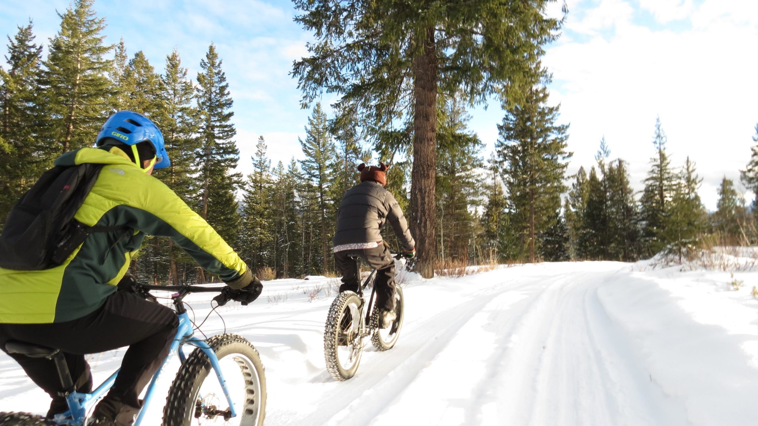 Two people biking on a snowy trail through a forested area. One cyclist is wearing a bright green and blue jacket with a helmet, while the other, seen from behind, is dressed in black and wearing a bear-themed hat. Tall evergreen trees surround the path under a partly cloudy sky. #2 and bottom of Razorback mountain bike trail.