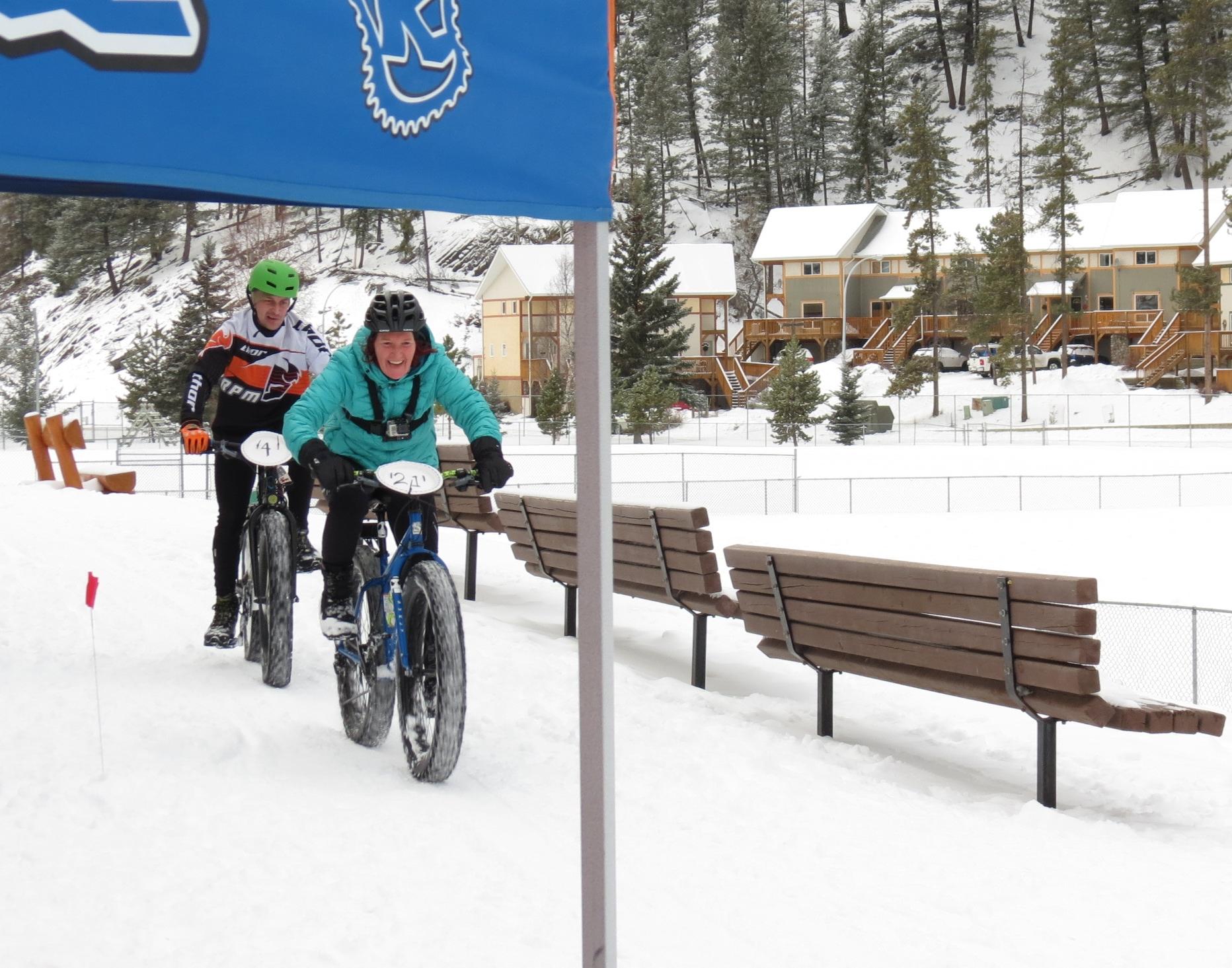 Two individuals riding fat tire bicycles through a snowy landscape, smiling and enjoying a winter biking event. In the background, wooden benches and snow-covered buildings are visible, surrounded by evergreen trees. Winter Cross mountain bike trail.