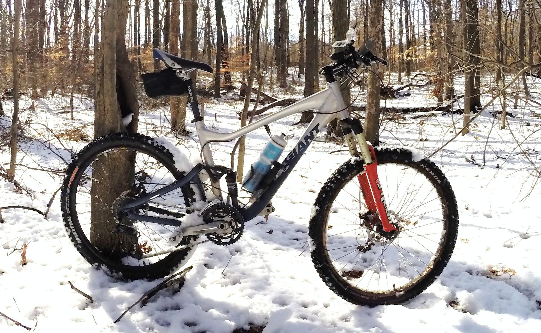 A mountain bike leaning against a tree in a snowy forest, with a partially obscured trail in the background. The bike features a silver frame, front suspension, and knobby tires, with a water bottle attached to the frame. The surrounding area is covered in a light layer of snow, and there are bare trees in the background. Wolfes Pond park mountain bike trail.