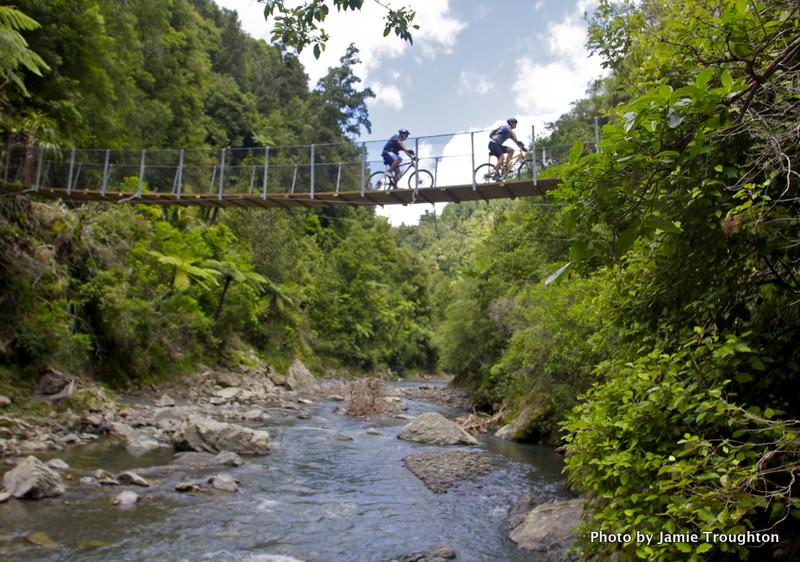 Two cyclists ride over a suspended wooden bridge surrounded by lush greenery, with a rocky river flowing below. The scene captures a bright day with blue skies and fluffy clouds. Motu Trails mountain bike trail.