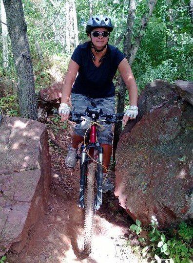 A cyclist navigating a rocky trail in a wooded area, wearing a helmet and sunglasses. The bike is positioned between two large rocks, surrounded by greenery and sunlight filtering through the trees. Cuyuna Lakes mountain bike trail.