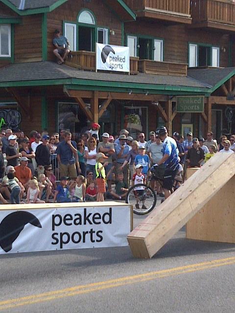 A crowd of spectators gathered in front of a wooden ramp, watching a cyclist preparing to perform a trick. The scene is set in a lively outdoor area with a building displaying a "Peaked Sports" sign in the background. People of various ages, including children, are closely observing the event, showing excitement and anticipation.