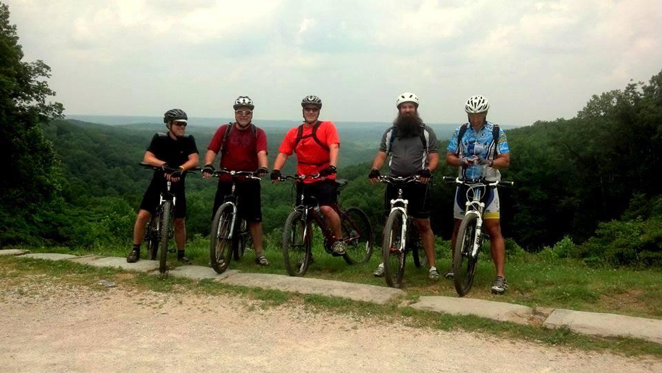 Five mountain bikers pose for a photo on a scenic overlook, surrounded by lush green hills and a cloudy sky. They are equipped with helmets and riding gear, standing beside their bikes on a gravel path. The atmosphere suggests a day of outdoor adventure and camaraderie. Brown County Park mountain bike trail.