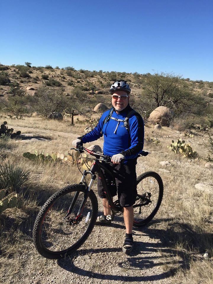 A mountain biker in a blue long-sleeve shirt and black shorts stands next to a mountain bike on a dirt trail surrounded by cacti and rocky terrain under a clear blue sky. 50-year Trail / Golder Ranch mountain bike trail.