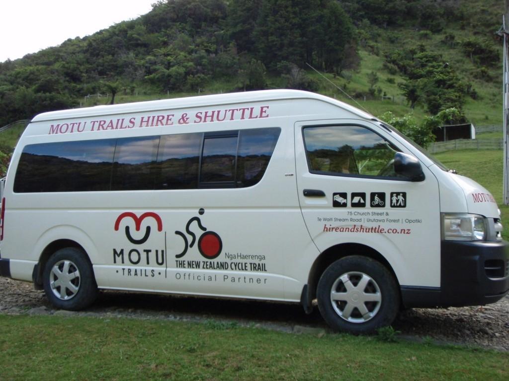 A white shuttle van with "MOTU TRAILS HIRE & SHUTTLE" prominently displayed on the side. The vehicle features icons representing cycling, walking, and other outdoor activities, along with contact information and the logo of the New Zealand Cycle Trail. It is parked on a gravel surface, surrounded by greenery and hills in the background. Motu Trails mountain bike trail.