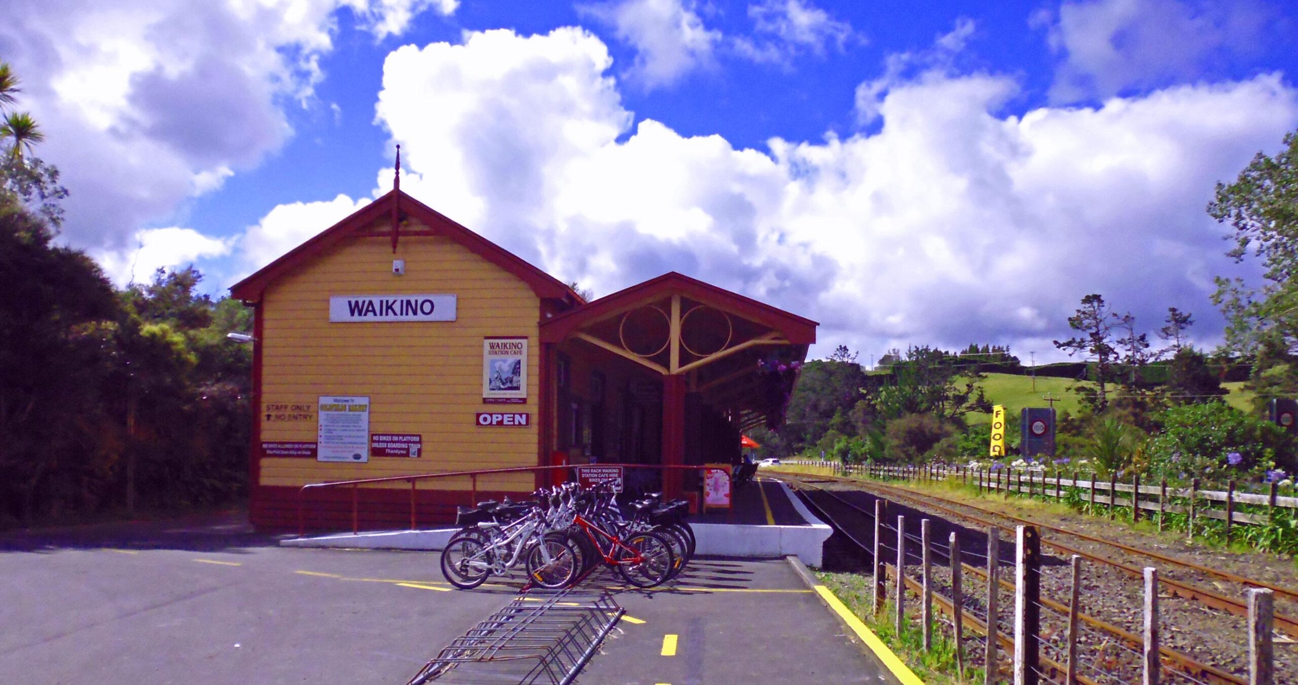 A brightly colored wooden train station labeled "Waikino" with a canopied platform. Bicycles are parked in the foreground, and the scene features lush greenery and a blue sky filled with clouds in the background. Hauraki Rail Trail mountain bike trail.