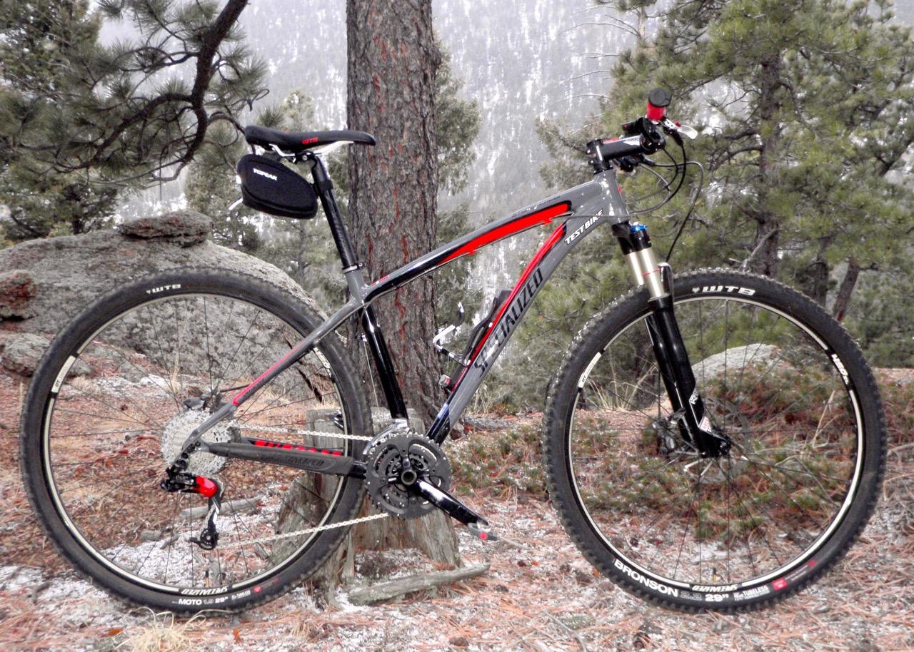 Specialized Stumpjumper Comp 29er: A mountain bike leaning against a tree in a forested area, featuring a gray and red frame, front suspension, and knobby tires. The background includes rocky terrain and pine trees.