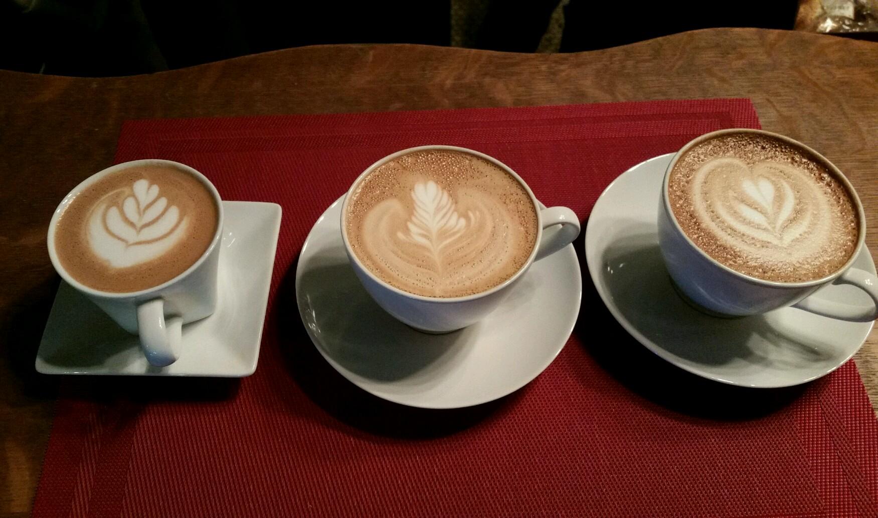 Three cups of coffee with intricate latte art, including designs of leaves and hearts, placed on white saucers atop a textured red table mat. The cups vary in size and style, adding a visually appealing arrangement.