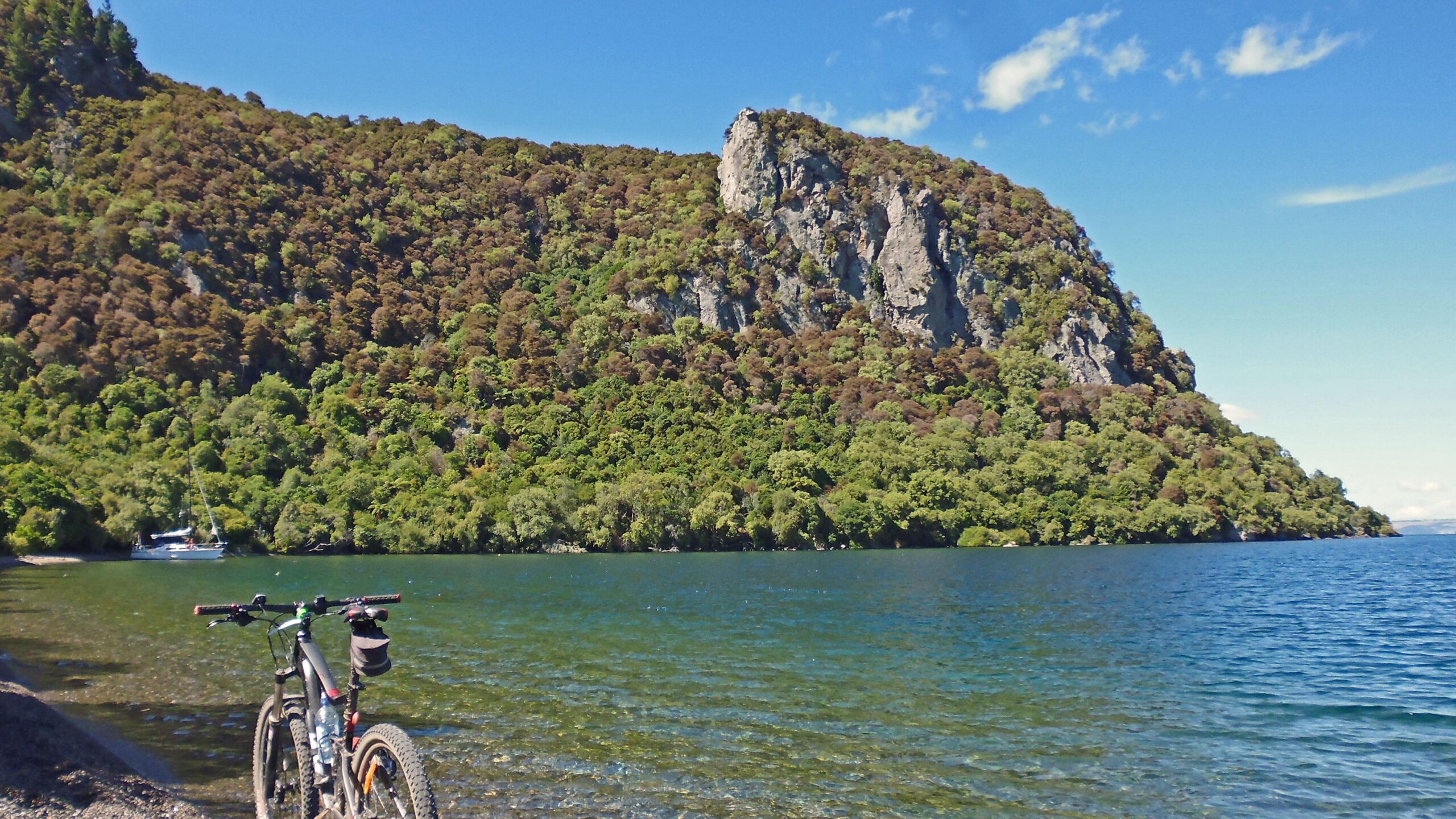 A scenic view of a lakeside landscape featuring a rocky hill covered in greenery. In the foreground, a mountain bike leans against a stony shoreline, while a calm, blue lake reflects the sky and surrounding trees. A sailboat can be seen in the background, further enhancing the tranquil outdoor setting. Great Lake Trails mountain bike trail.