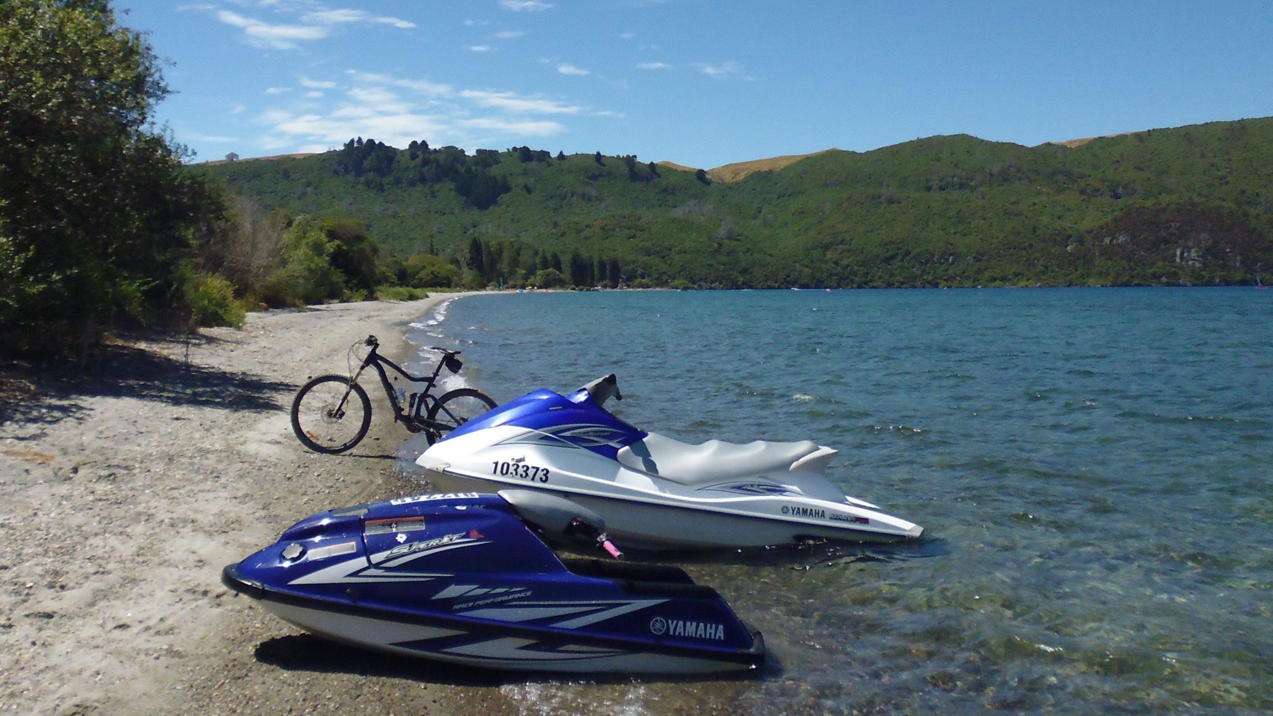 Two jet skis are positioned on a sandy shore beside a calm lake, with a bicycle leaning against the shore. The backdrop features lush green hills under a clear blue sky with scattered clouds, creating a serene outdoor scene. Great Lake Trails mountain bike trail.