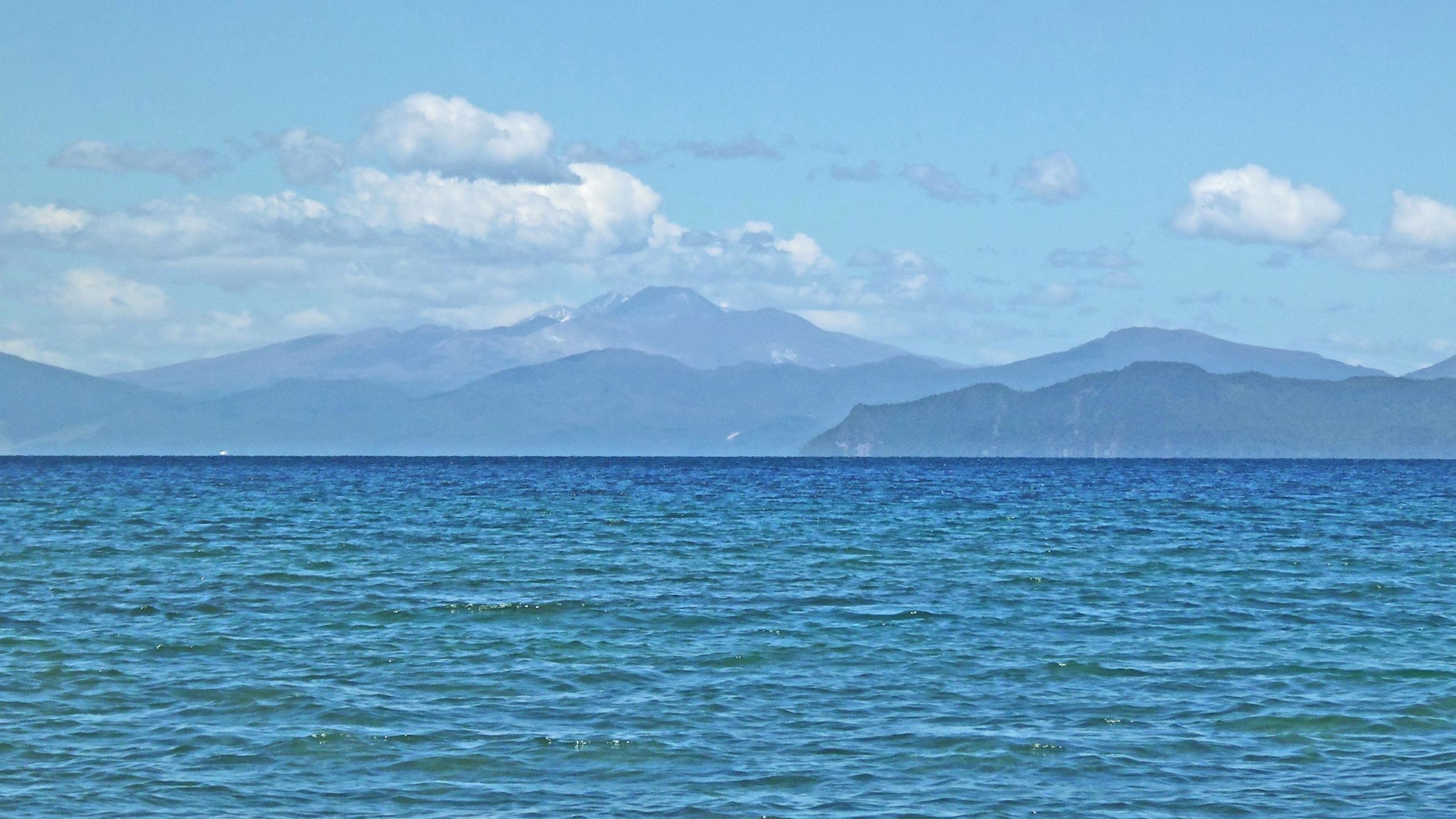 A serene view of the ocean with gentle waves, stretching towards distant mountains under a clear blue sky dotted with fluffy white clouds. Great Lake Trails mountain bike trail.