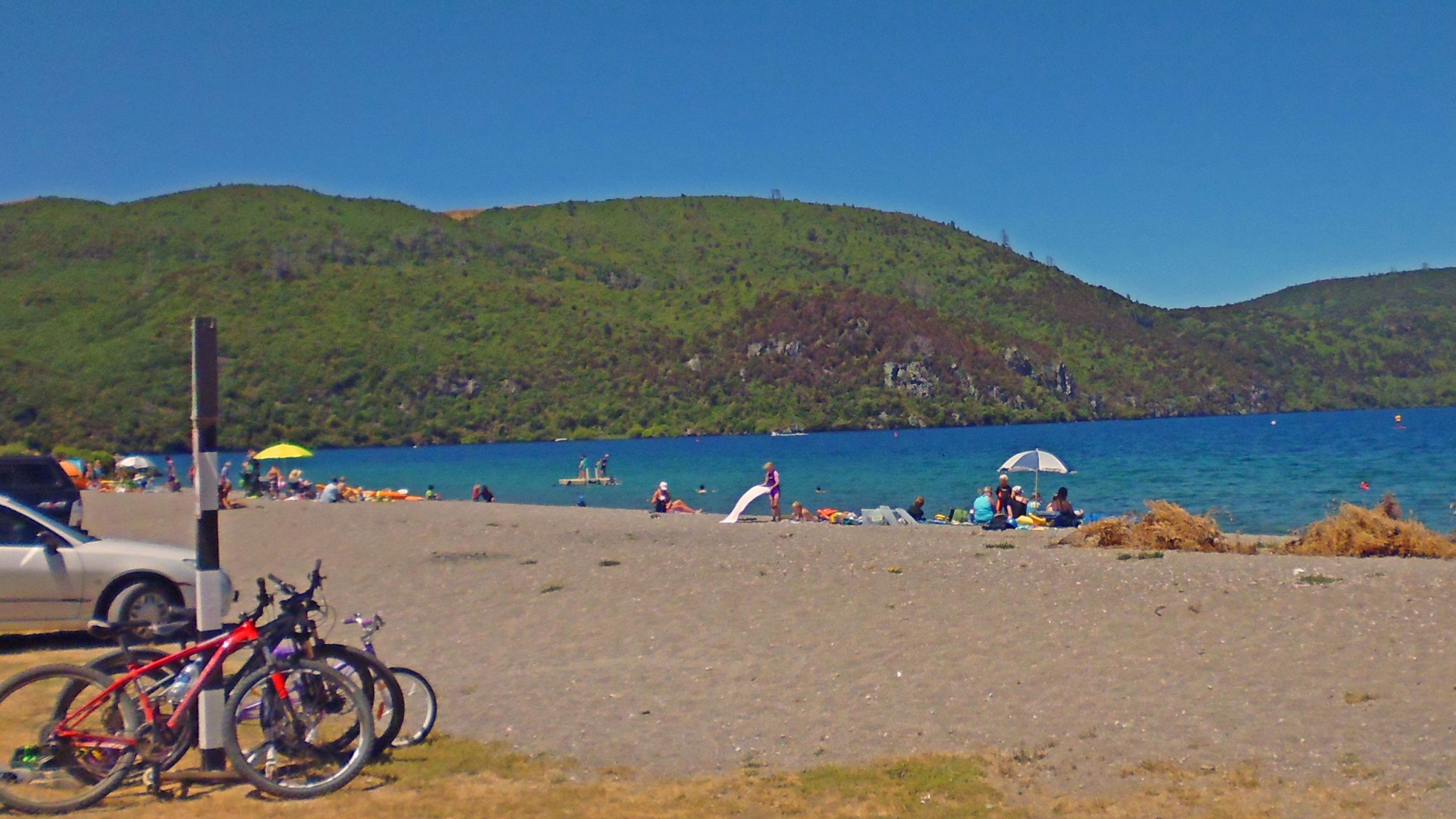 A scenic view of a beach with a sandy shore and blue water, set against a backdrop of green hills. People are relaxing on the beach, some are sitting under umbrellas, while others are in the water or on a dock. Bicycles are parked nearby, and a few cars are visible in the foreground. The sky is clear and sunny, creating a vibrant and inviting atmosphere. Great Lake Trails mountain bike trail.