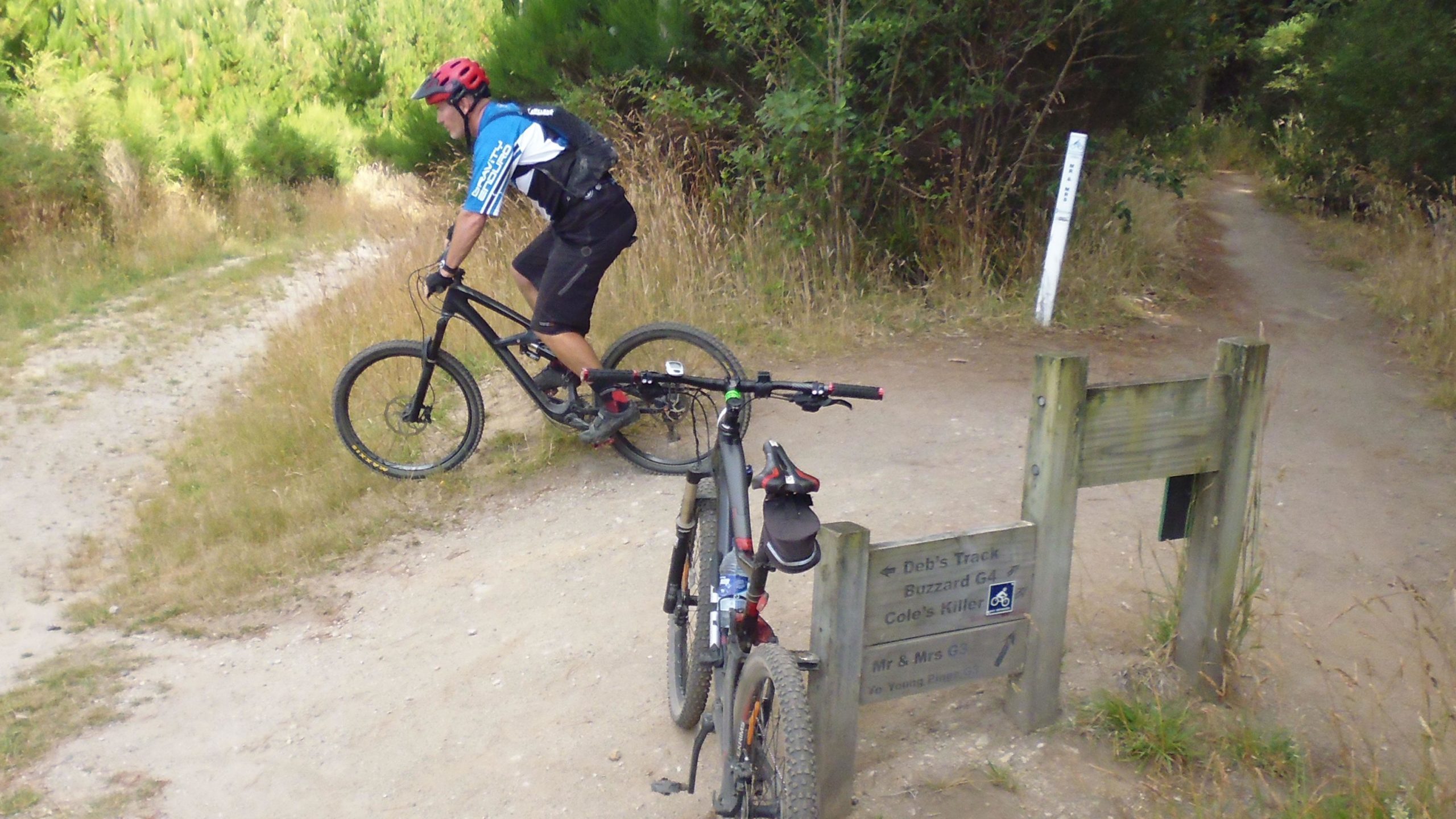 A mountain biker pauses on a gravel path surrounded by lush greenery, with two bicycles visible: one is parked next to a wooden signpost indicating trail names such as "Deb's Track" and "Buzzard G4." The cyclist is wearing a helmet and a blue jersey, while the path diverges into two directions. Craters Mountain Bike Park mountain bike trail.