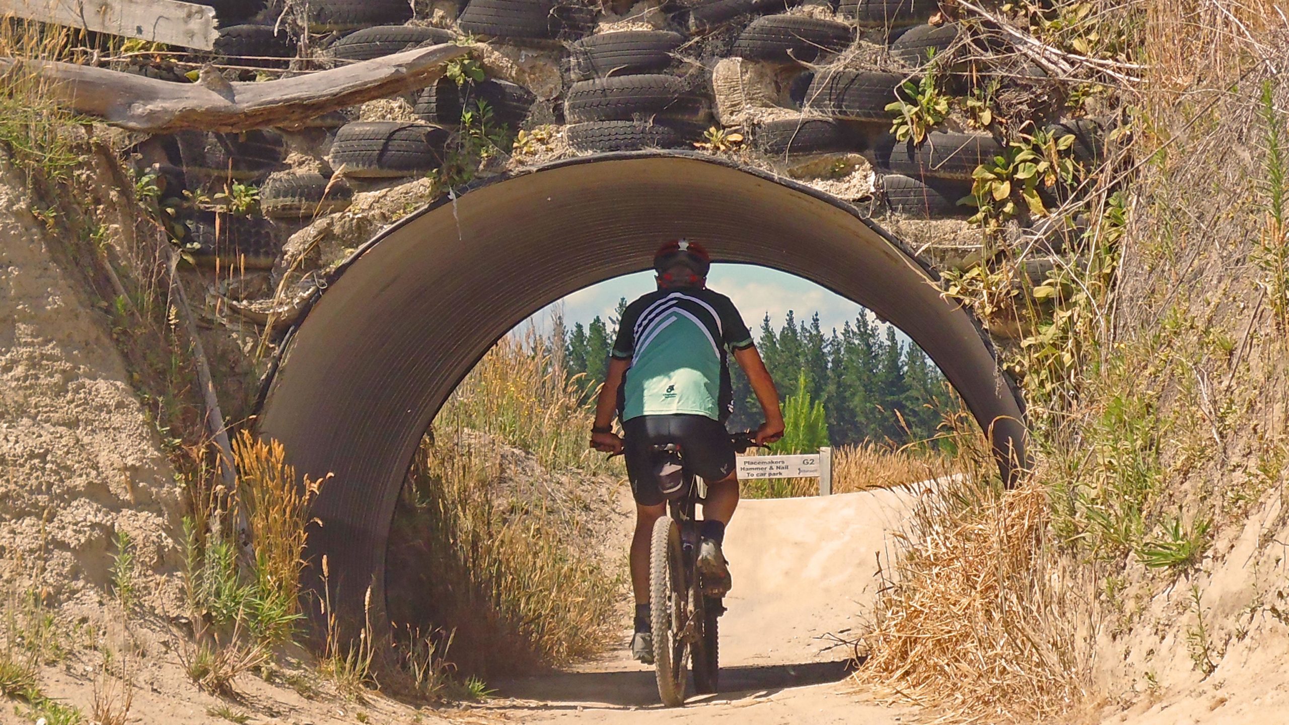 A cyclist riding a mountain bike through a tunnel made of metal piping, surrounded by tall grass and overgrown vegetation. The tunnel is built into a sandy path, with a backdrop of trees visible in the distance. A small sign can be seen near the tunnel entrance. Craters Mountain Bike Park mountain bike trail.