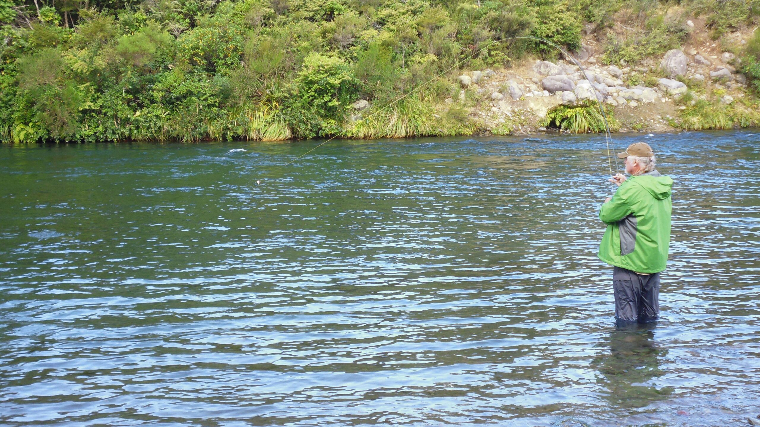 A person fishing in a river, wearing a green jacket and a cap, standing in the water surrounded by lush greenery and rocky terrain. The water is clear and reflects the natural surroundings. Tongariro River Trail mountain bike trail.