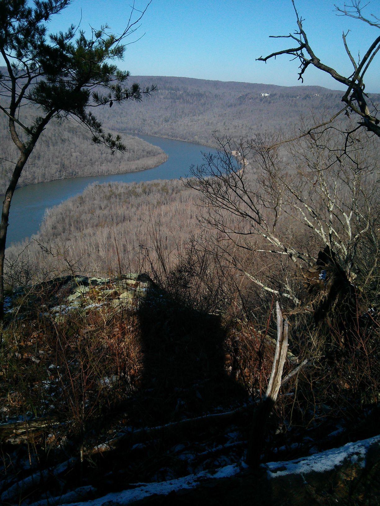 A scenic view from a high vantage point overlooking a winding river surrounded by a landscape of bare trees, under clear blue skies. Shadows cast by nearby vegetation and rock formations add depth to the foreground. Raccoon Mountain Trail Network mountain bike trail.