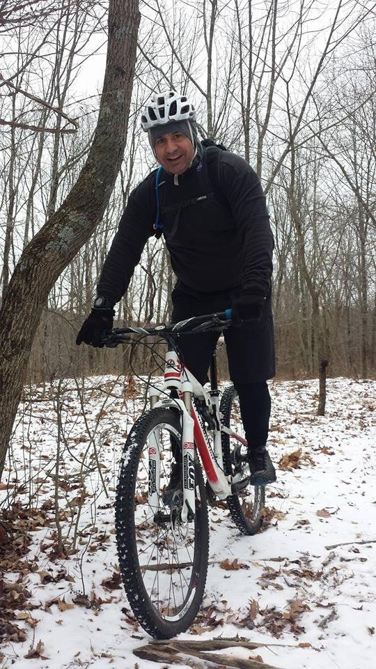 A person on a mountain bike smiles while standing in a snowy forested area. The cyclist is wearing a helmet, gloves, and winter clothing, with sparse trees and fallen leaves visible in the background. Snow covers the ground, indicating a cold winter day. Ringwood Skylands Manor mountain bike trail.