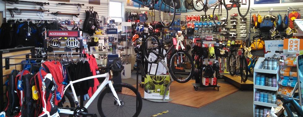 Interior of a bike shop displaying a variety of bicycles, cycling gear, and accessories. Shelves are stocked with helmets, jerseys, water bottles, and other cycling equipment, while bicycles are prominently featured throughout the room.
