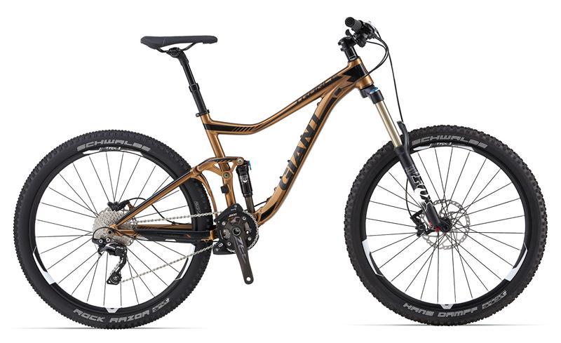 An image of a mountain bike with a bronze frame, featuring dual suspension, wide tires, and a modern design. The bike is equipped with a front suspension fork, disc brakes, and a Shimano gear system, showcasing its suitability for rugged terrains. The tires are labeled "Schwalbe Rock Razor" and "Hand Damping," indicating high performance for off-road cycling.
