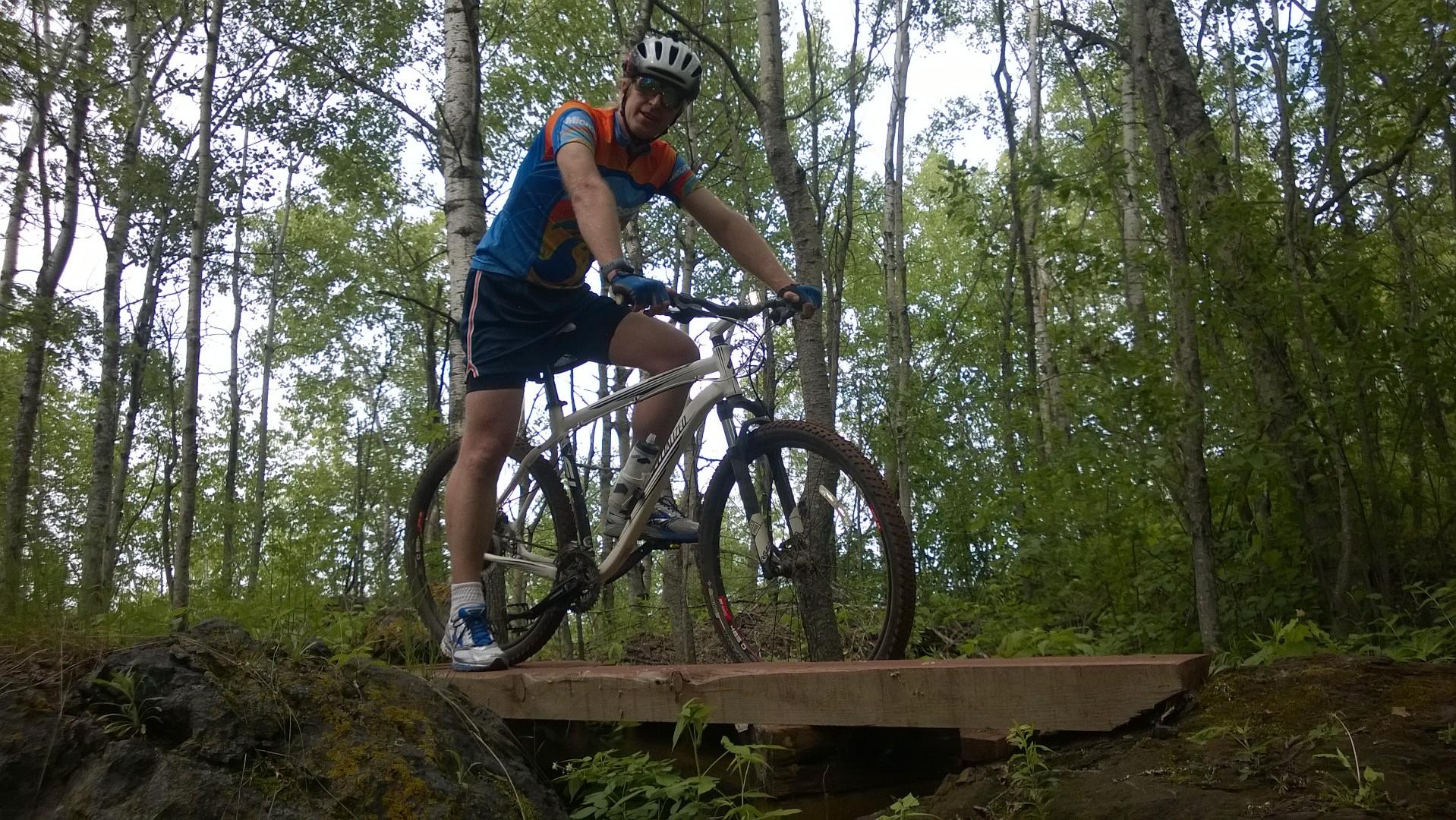 A mountain biker in a blue and orange jersey and helmet, standing on a wooden bridge in a forested area, surrounded by green trees. Cuyuna Lakes mountain bike trail.