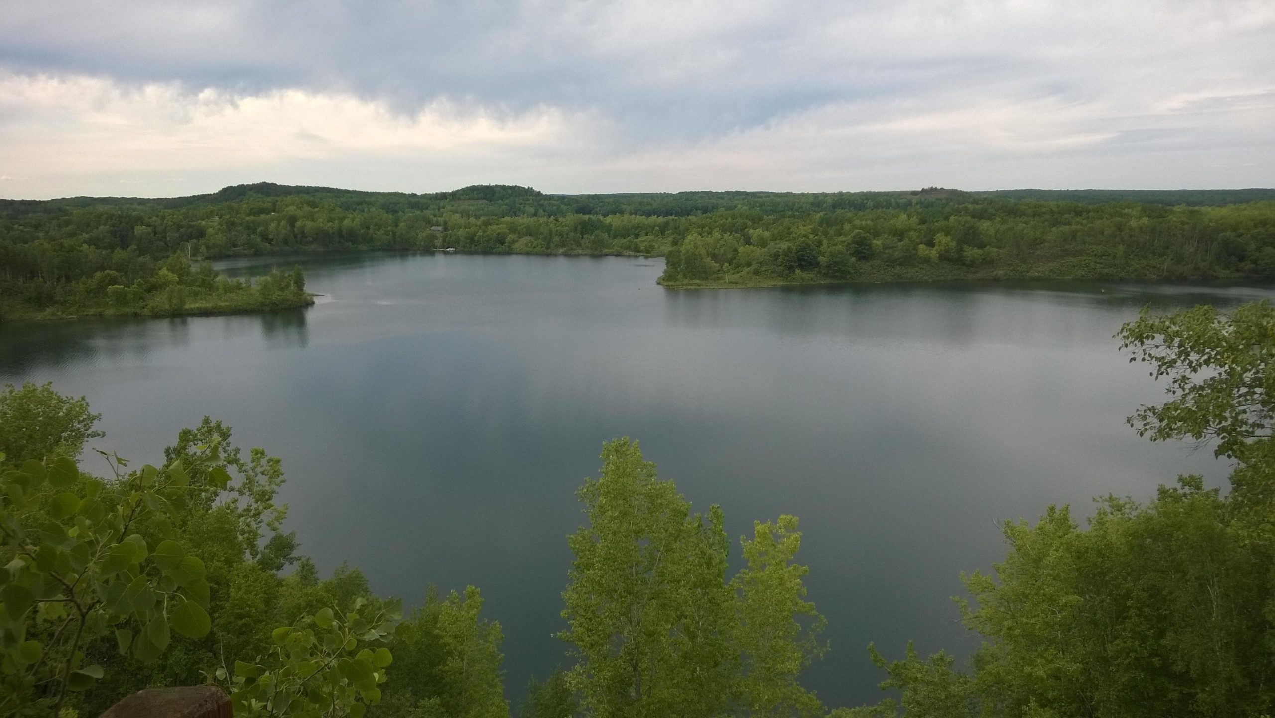 A serene landscape featuring a calm lake surrounded by lush greenery and rolling hills, under a cloudy sky. The water reflects the greenery, creating a peaceful and tranquil atmosphere. Cuyuna Lakes mountain bike trail.