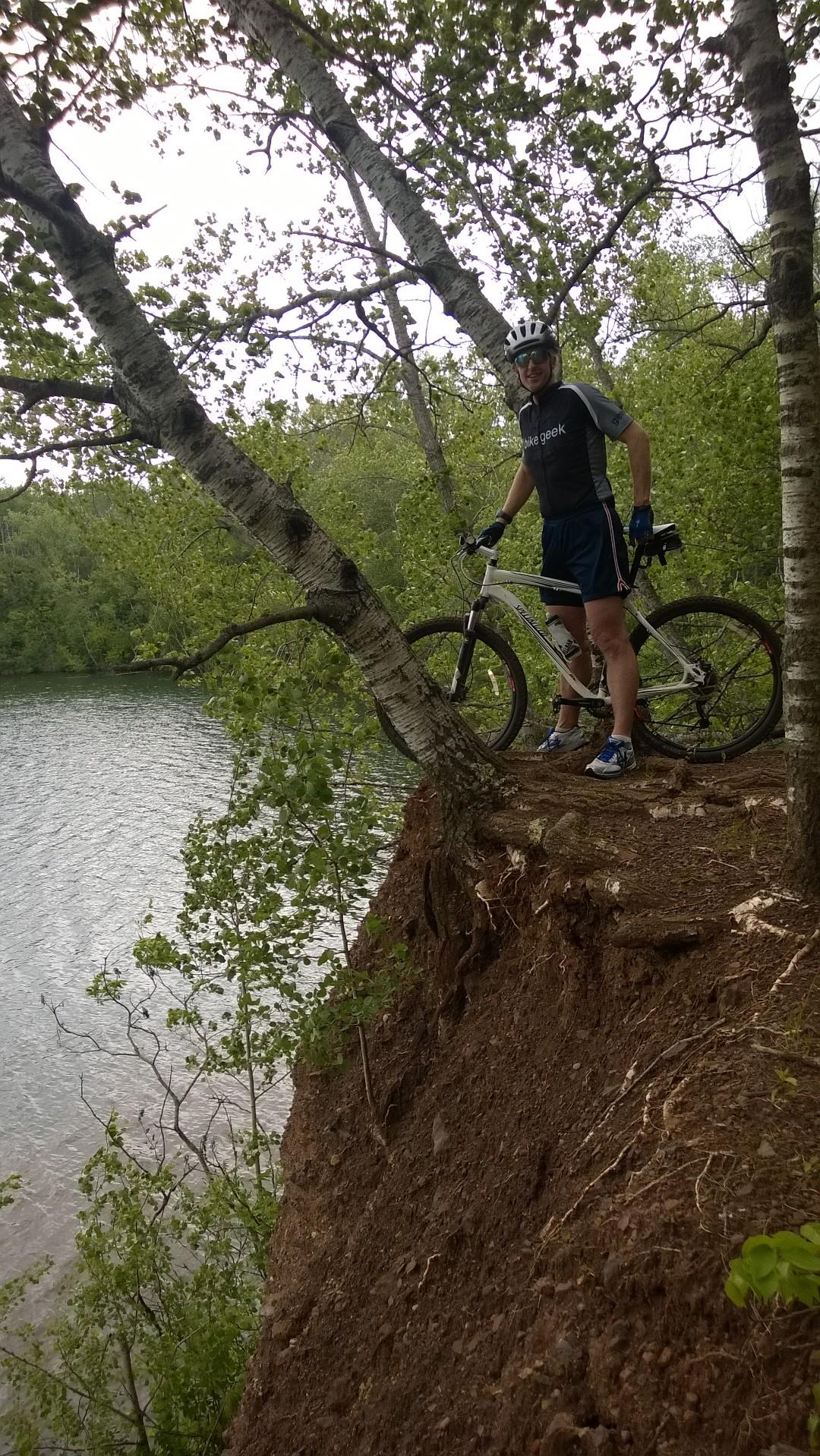 A mountain biker standing on the edge of a dirt slope near a lake, surrounded by green trees. The biker is wearing a helmet, sunglasses, and a shirt with the text "bike geek." The scene captures a lush, natural environment with water visible in the background. Cuyuna Lakes mountain bike trail.