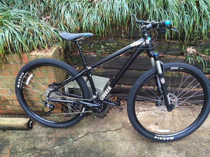 Charge Cooker 4: A black mountain bike leaning against a brick wall, with a gravel pathway visible in the foreground. The bicycle features a sturdy frame, wide tires with a textured tread, and a front suspension fork. Green foliage is in the background, adding a natural setting to the scene.
