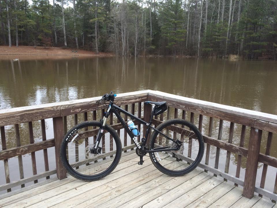 Charge Cooker 4: A mountain bike leaning against a wooden railing by a calm lake, surrounded by trees. The bike has a water bottle attached and is positioned on a wooden deck, with reflections in the water visible in the background.