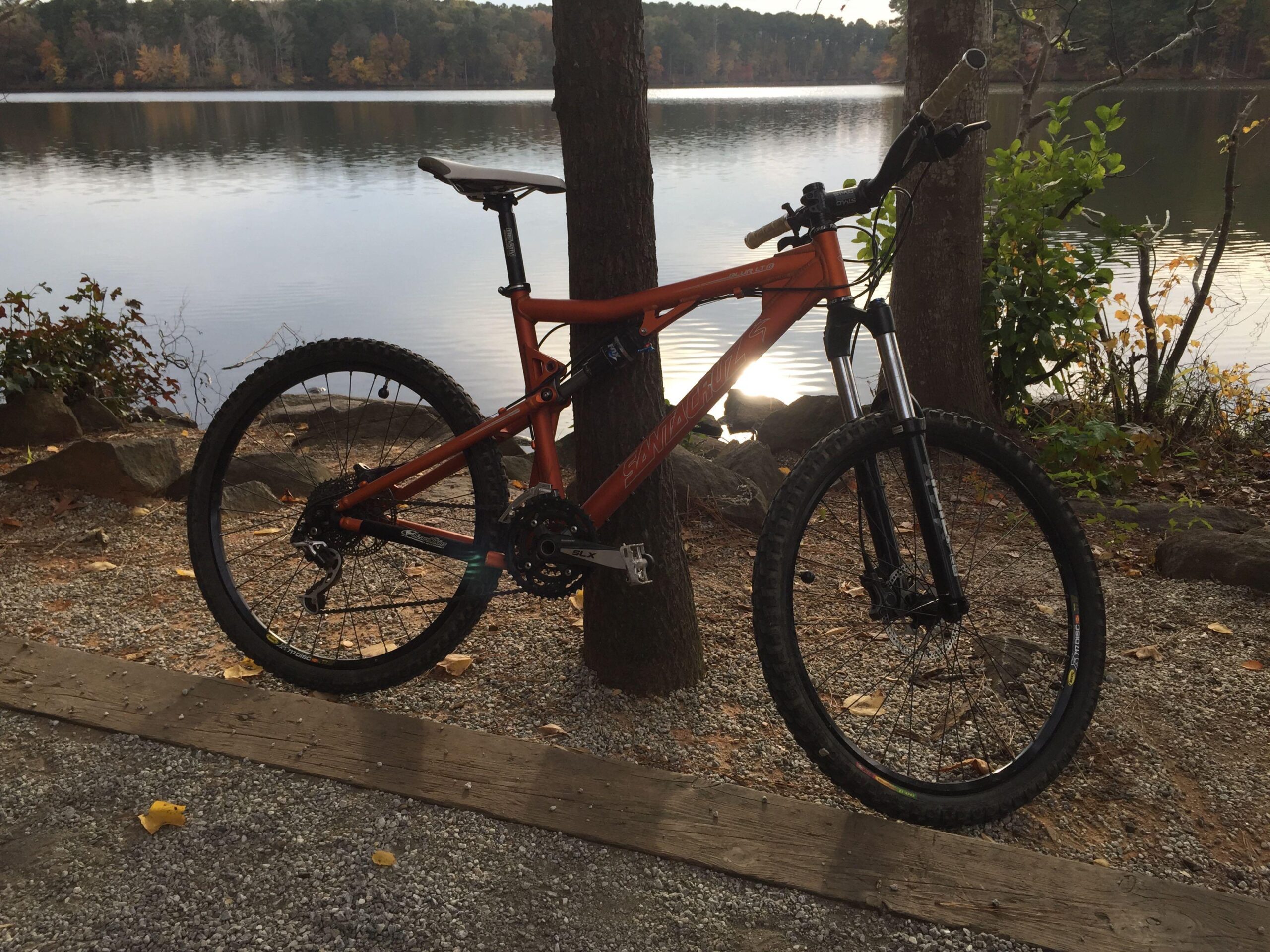 Santa Cruz Blur LT: An orange mountain bike leaning against a tree near a calm lake, surrounded by rocky terrain and autumn foliage. The water reflects the trees and sky, creating a serene outdoor scene.