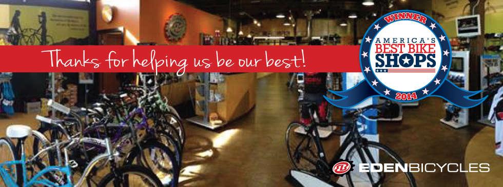 Interior view of a bicycle shop featuring a variety of bikes lined up for display. The shop has warm lighting and a welcoming atmosphere. There is a banner that reads, "Thanks for helping us be our best!" along with a badge indicating the shop was recognized as a winner in "America's Best Bike Shops 2014." The shop is branded with the "Eden Bicycles" logo.