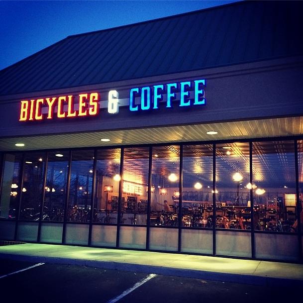 A storefront with large illuminated signs reading "BICYCLES & COFFEE." The front features large windows showcasing bicycles inside, with warm lighting visible. The image is taken at dusk, giving a cozy atmosphere to the scene.