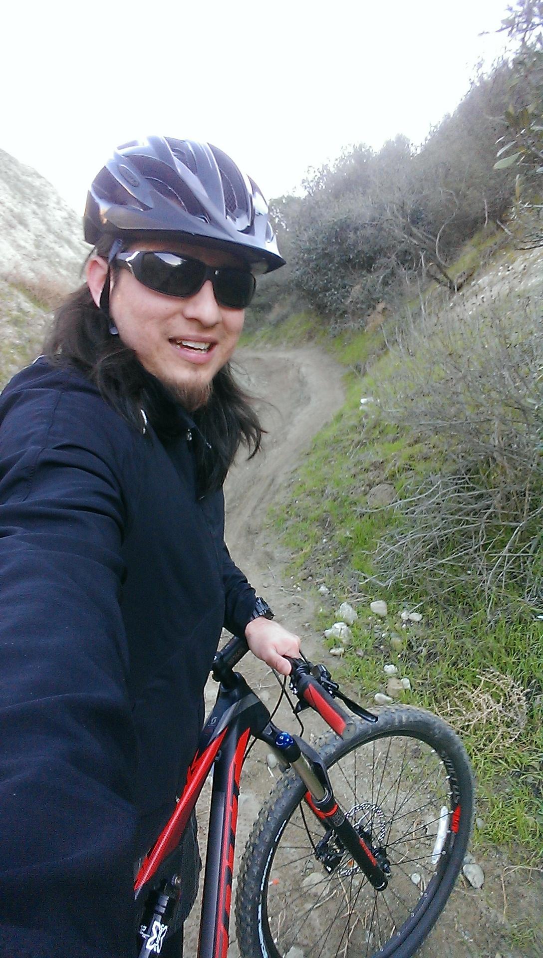 Scott Scott Spark 760: A person with long hair wearing a black helmet and sunglasses is smiling while holding a red mountain bike. They are standing on a dirt path surrounded by green vegetation and trees.