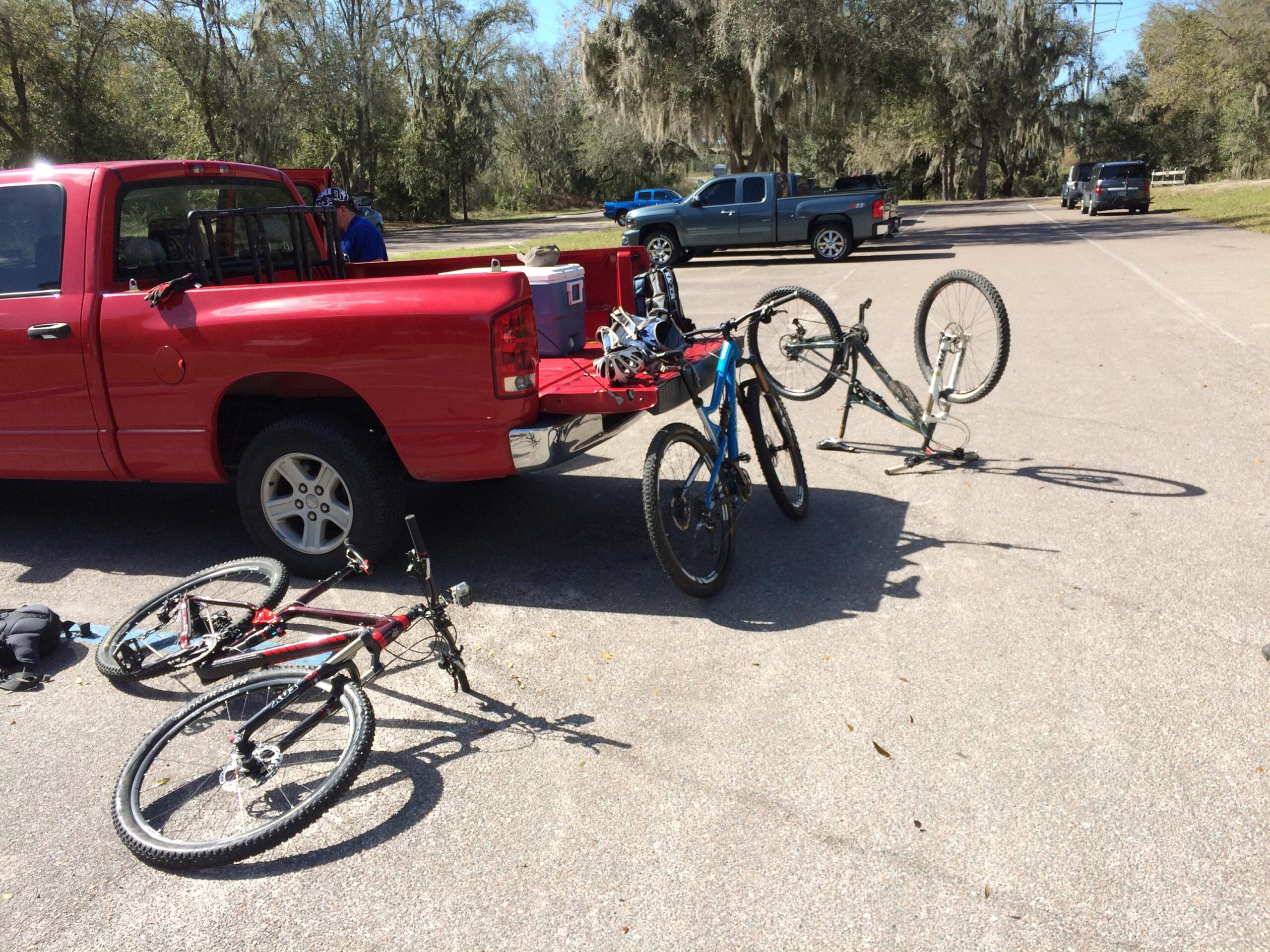A red pickup truck parked in a lot, with several bicycles lying on the ground beside it. One bike is upright with its front wheel in the air, while two others are lying on their sides. A person is seen in the truck bed, appearing to be organizing the cargo. The surrounding area features trees and other parked vehicles in the background. Alafia River State Park mountain bike trail.