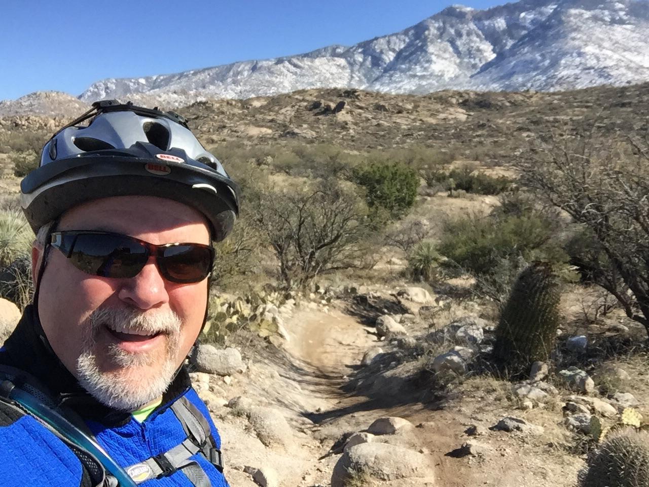 A smiling person wearing a blue jacket, sunglasses, and a helmet poses for a selfie on a rocky biking path. In the background, a mountain range is visible under a clear blue sky, surrounded by desert vegetation including cacti and shrubs. 50-year Trail / Golder Ranch mountain bike trail.