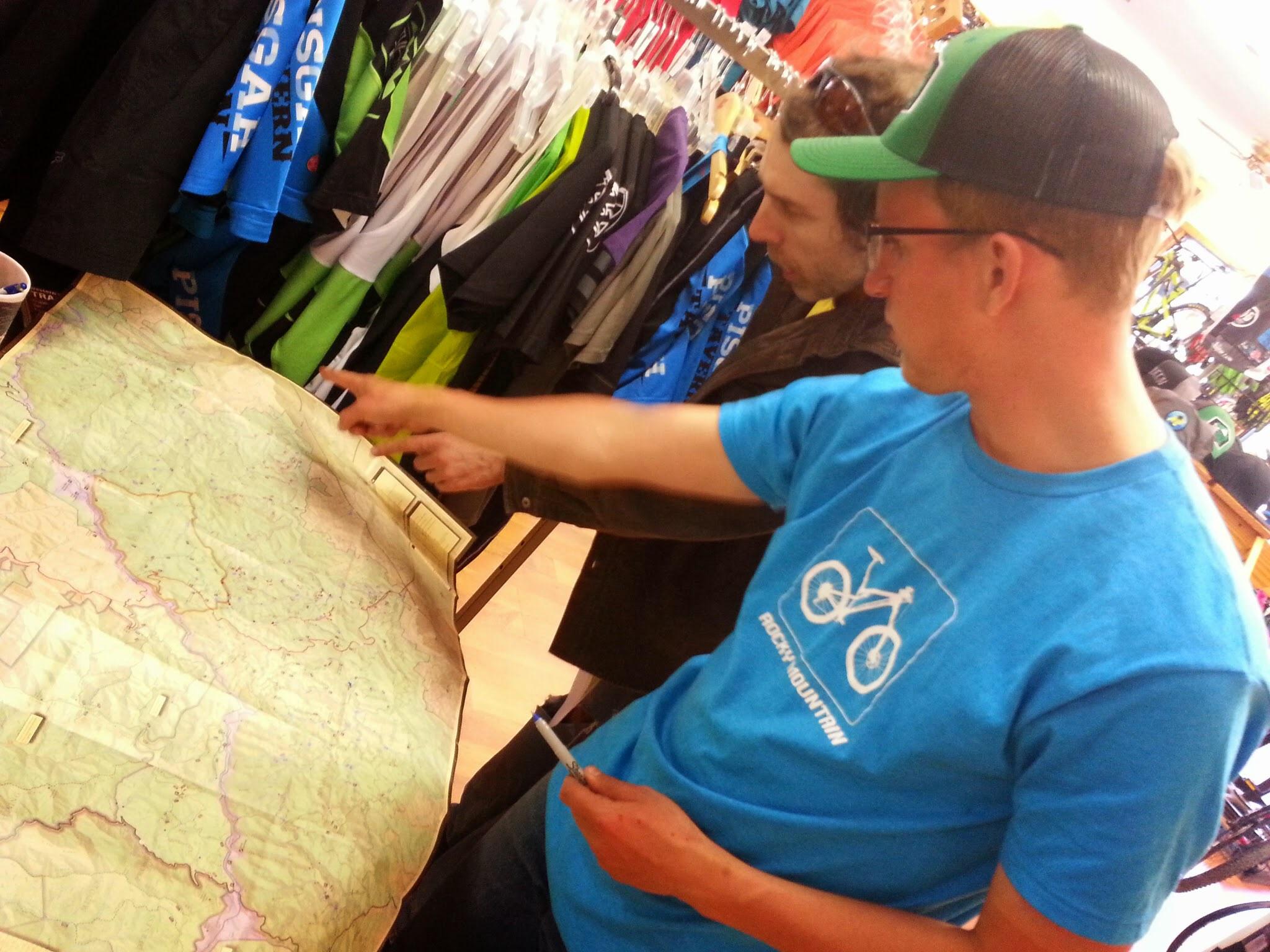 Two men are closely examining a large map laid out on a table in a sporting goods store. One man, wearing a blue t-shirt with a bicycle graphic, points to a specific area on the map, while the other man, dressed in a dark jacket and hat, looks on attentively. A variety of colorful cycling apparel hangs in the background.