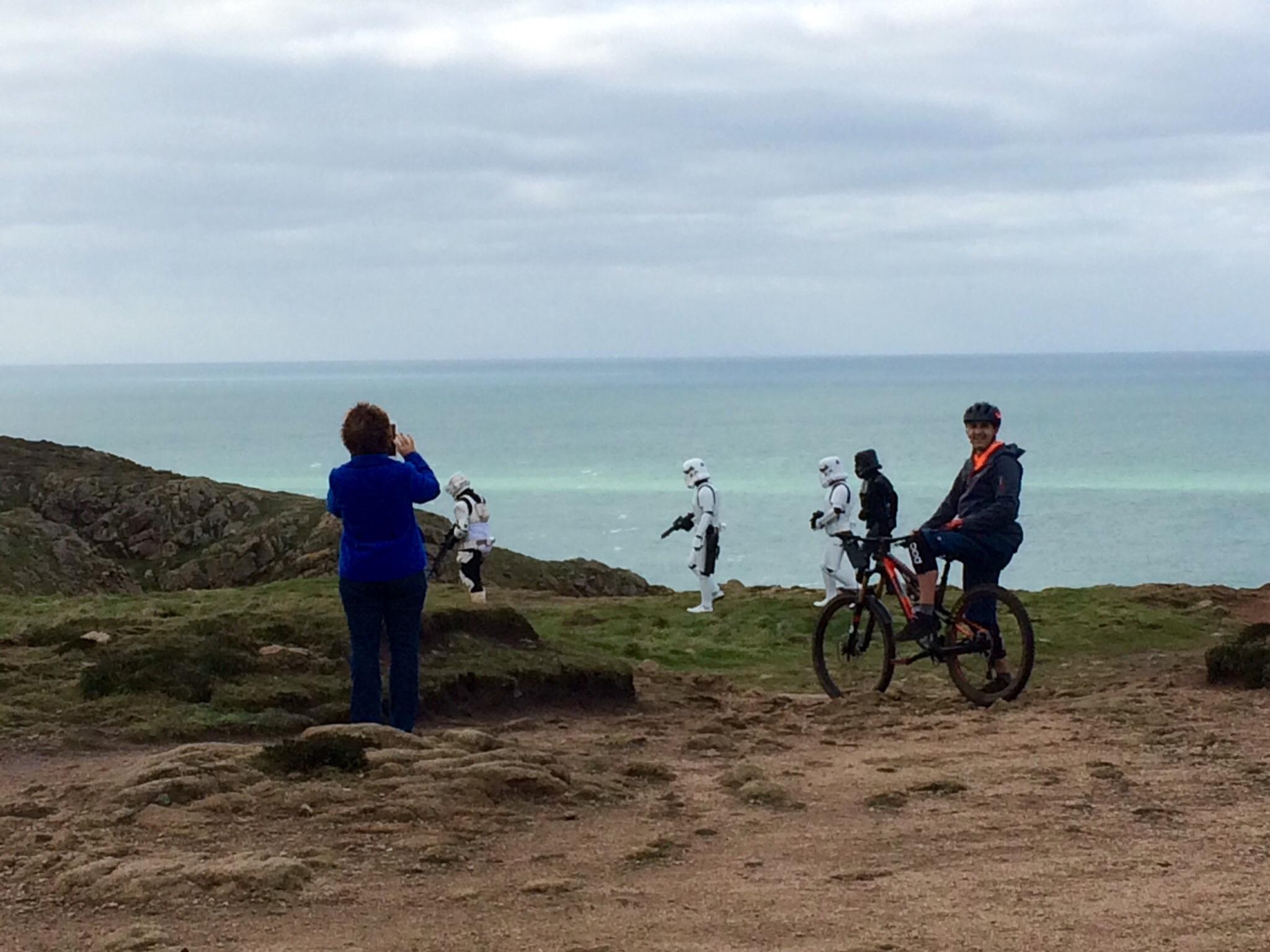 A person wearing a blue jacket takes a photo with a camera while standing on a grassy area near the coast. In the background, two figures dressed in stormtrooper costumes walk along the shoreline, while a person on a mountain bike, wearing a dark jacket with an orange accent, smiles at the camera. The scene features rocky terrain and a cloudy sky over a turquoise sea. #Allthelads mountain bike trail.