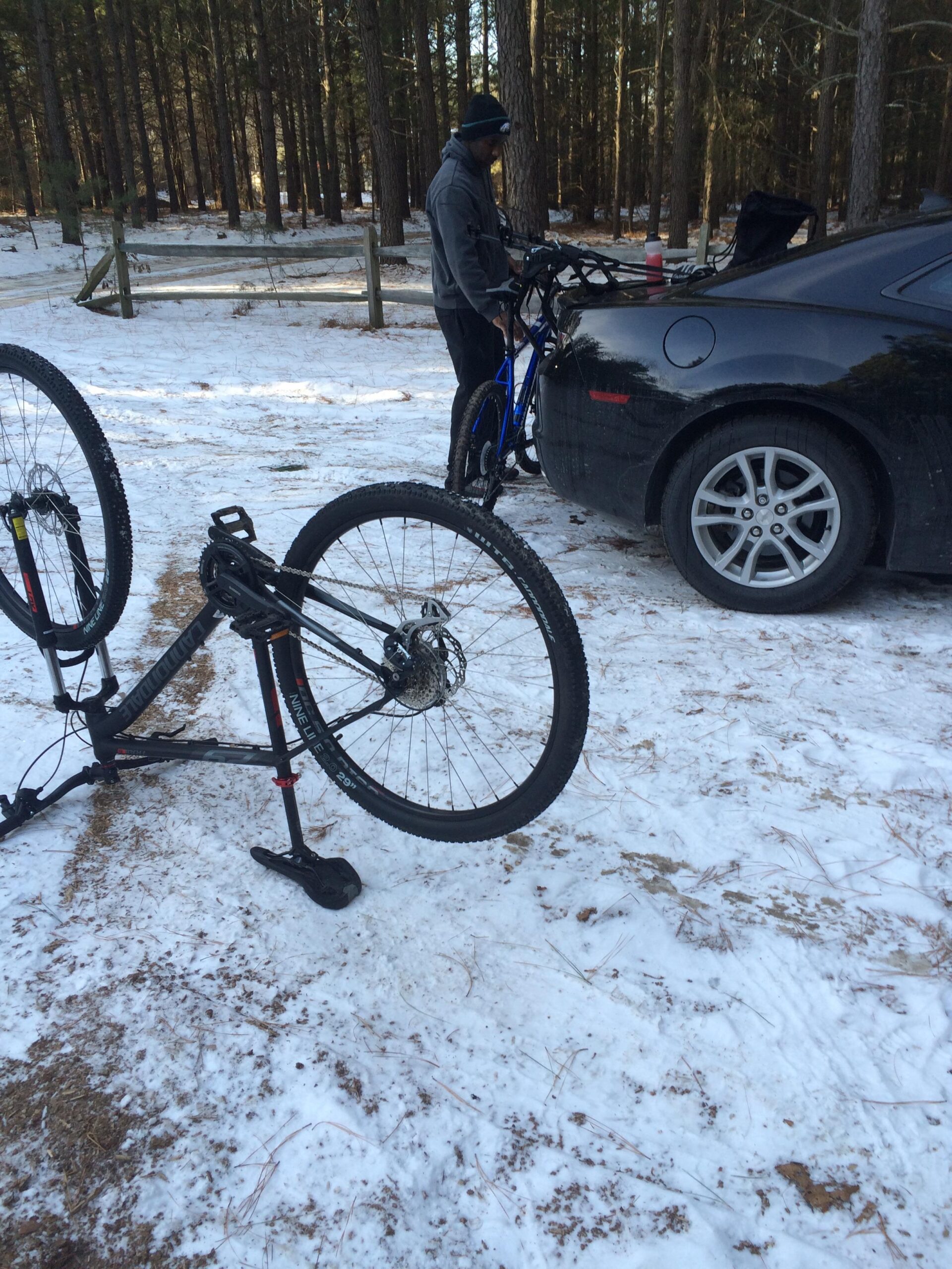 A person standing near a car in a snowy area, adjusting a blue bicycle. A second bicycle is positioned on its side in the foreground, with snow-covered ground and pine trees in the background. The bluffs mountain bike trail.