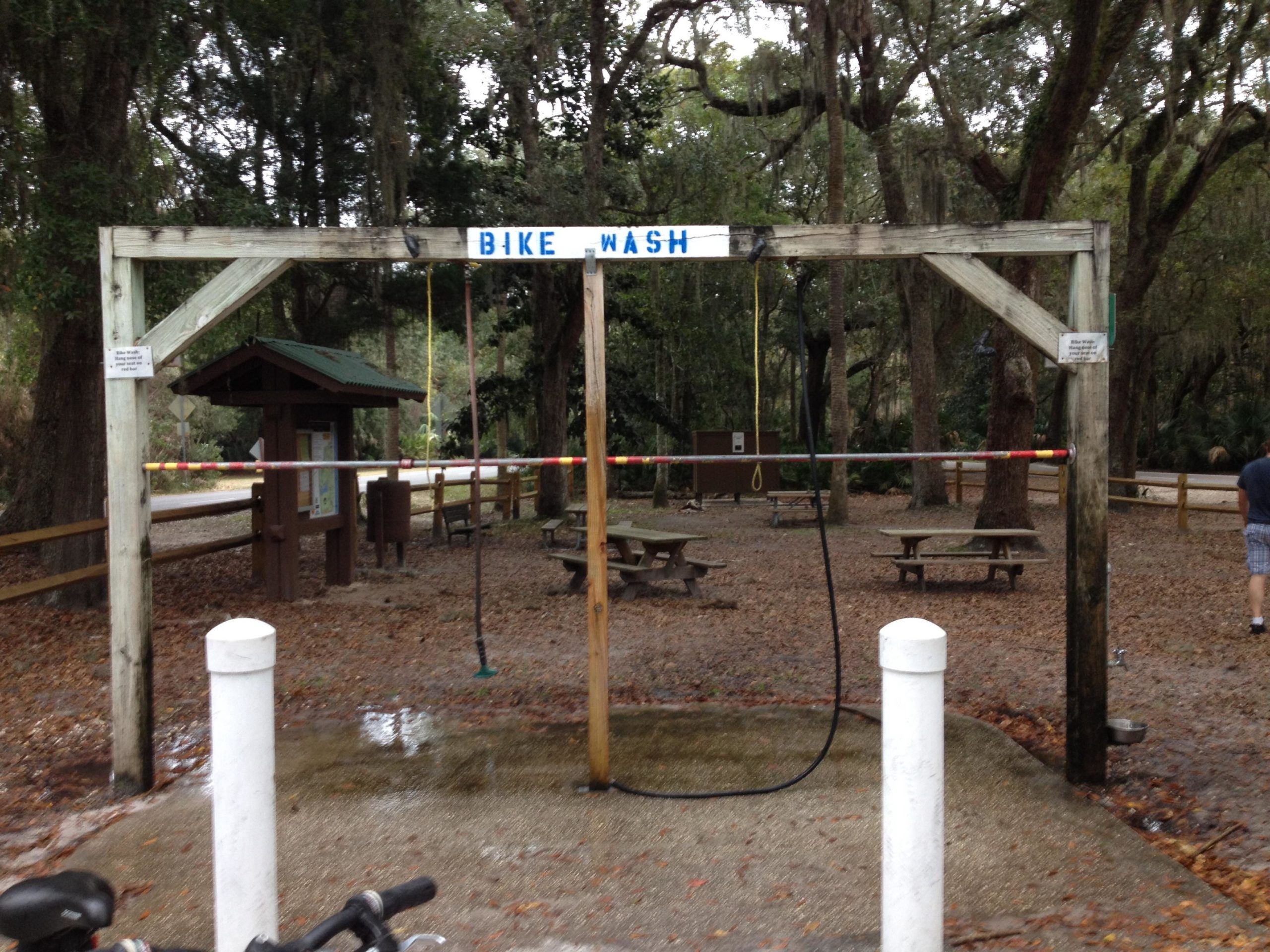 Wooden structure labeled "BIKE WASH" with hoses and a spray nozzle set in a nature area. Picnic tables and informational signage are visible in the background, surrounded by trees and fallen leaves. Kathryn Abby Hanna Park mountain bike trail.
