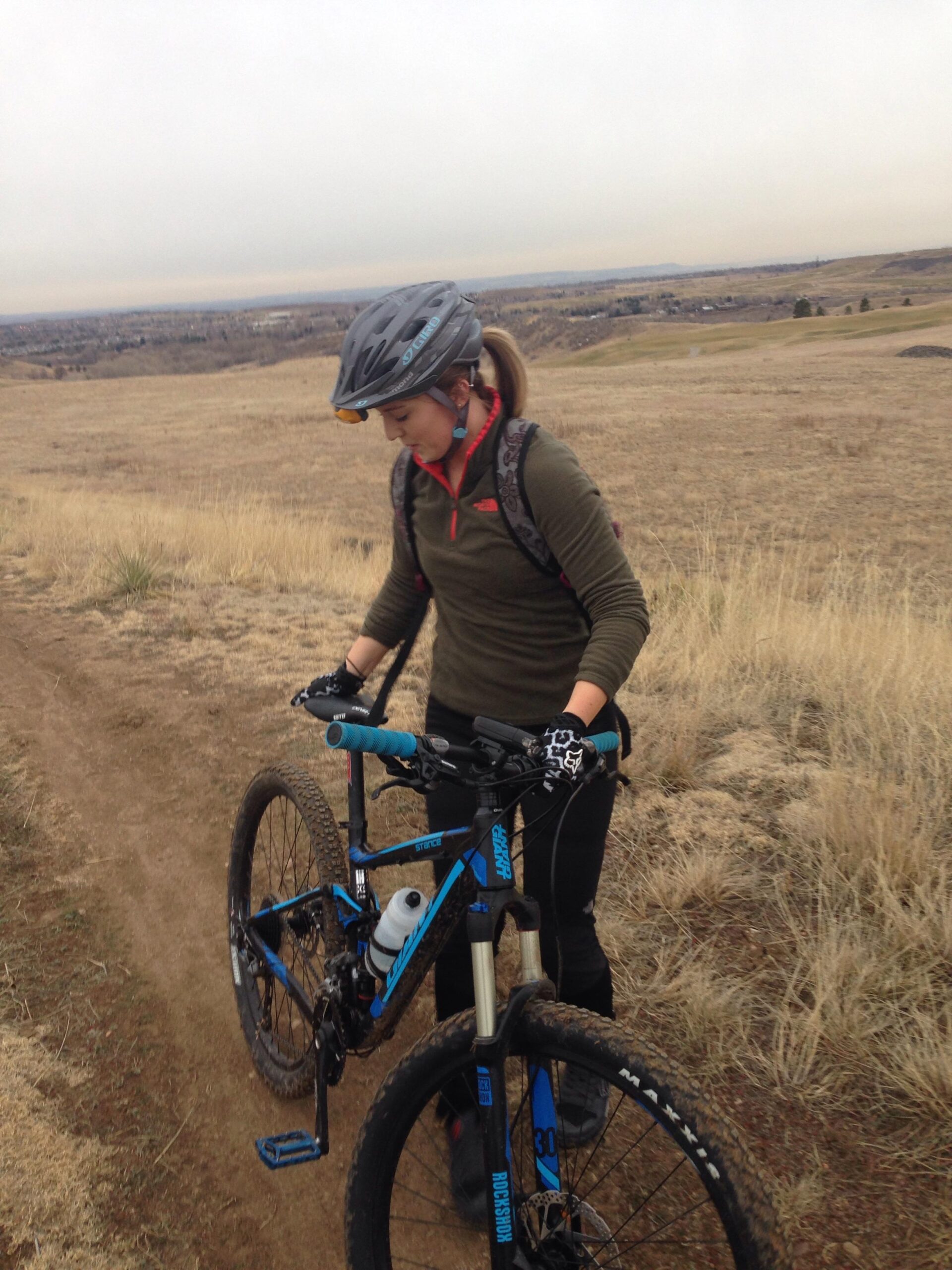 Giant Giant Stance 2: A person wearing a helmet and gloves stands beside a mountain bike on a dirt trail. They are looking down at the bike, with grassy terrain and a cloudy sky in the background. The landscape appears hilly and open, typical of a rural outdoor setting.