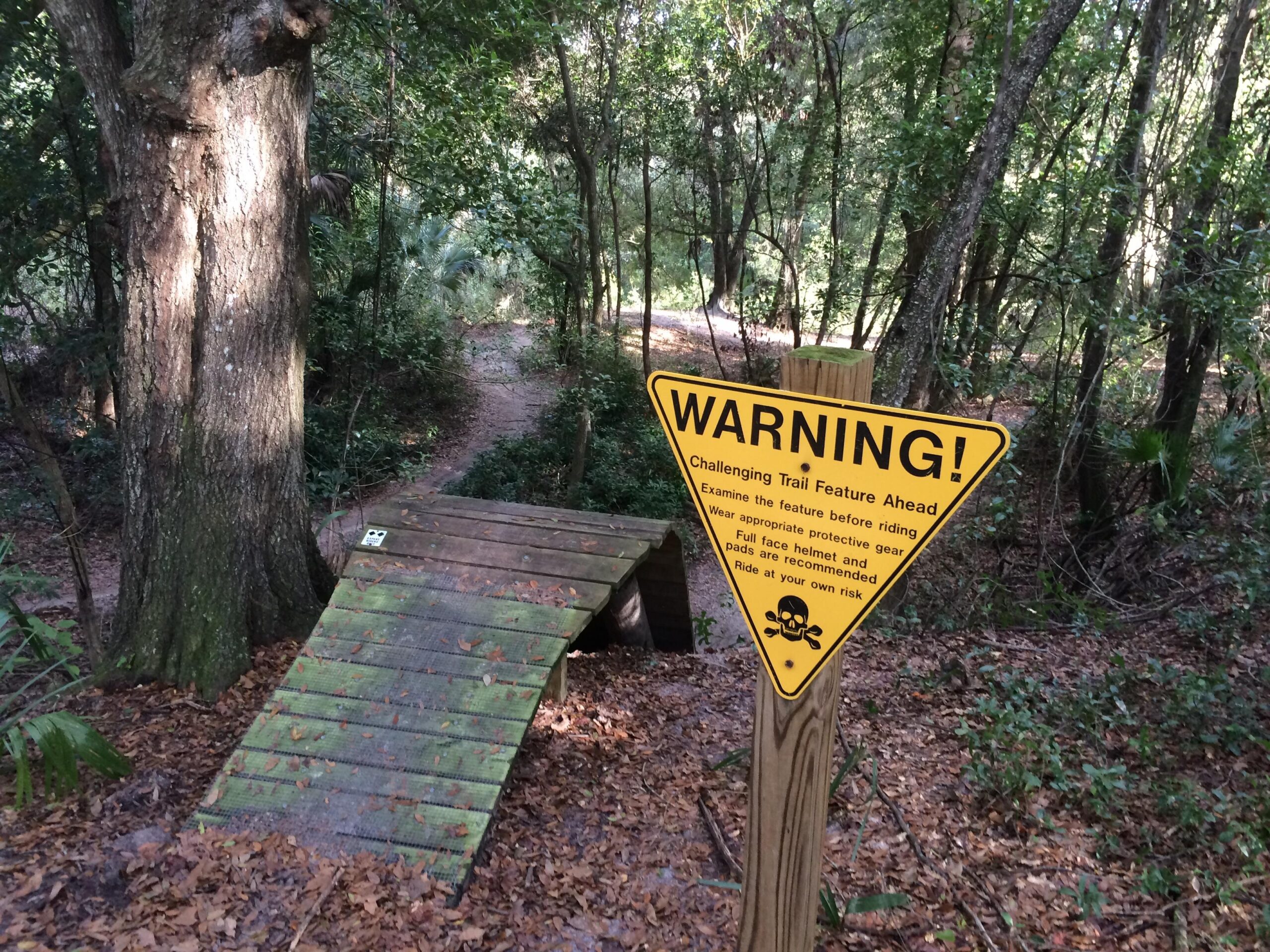 A wooden ramp with a textured surface leads down a wooded trail, surrounded by trees and foliage. A bright yellow warning sign stands nearby, alerting users to a challenging trail feature ahead and advising them to examine the ramp, wear protective gear, and ride at their own risk. The ground is covered in fallen leaves. Mount Dora Trail mountain bike trail.