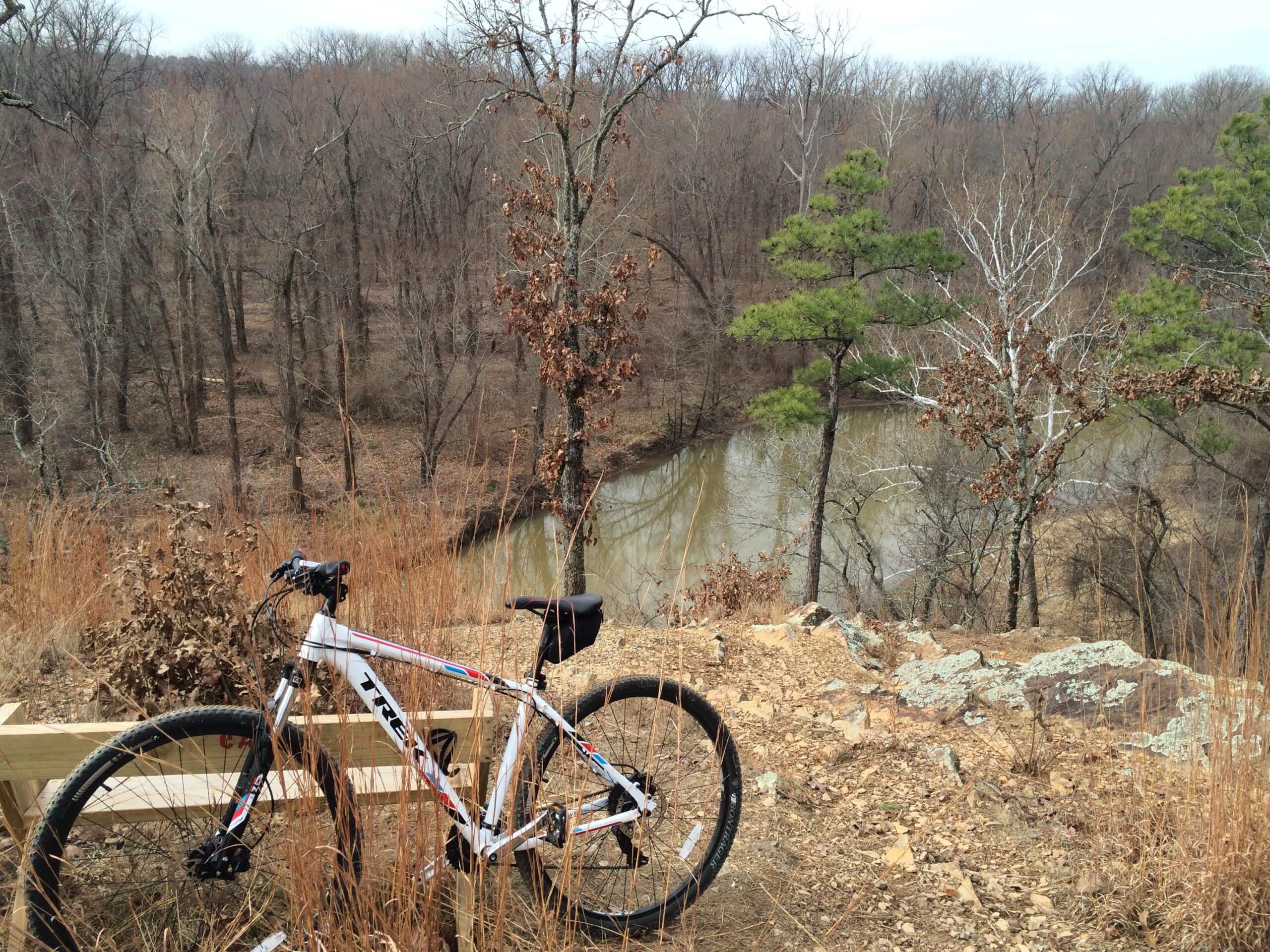 A mountain bike rests on a wooden bench overlooking a scenic view of a river and bare trees in a wooded area during early spring. The landscape features rocky terrain and patches of dry grass, with a few green pine trees contrasting against the brown foliage. The sky is overcast, adding a serene atmosphere to the outdoor scene. Burns Park mountain bike trail.