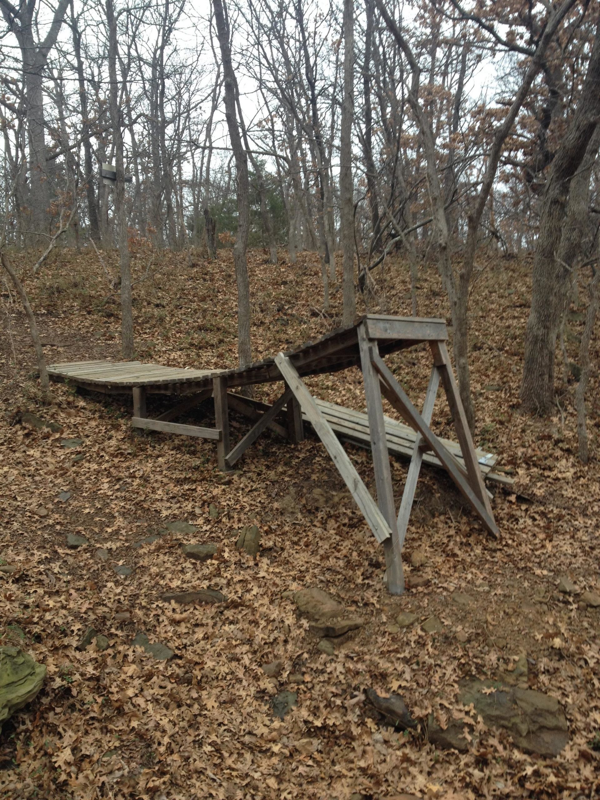 A wooden structure partially collapsed on a forest floor covered with fallen leaves. The surrounding area features bare trees and a sloping hillside. Turkey Mountain mountain bike trail.