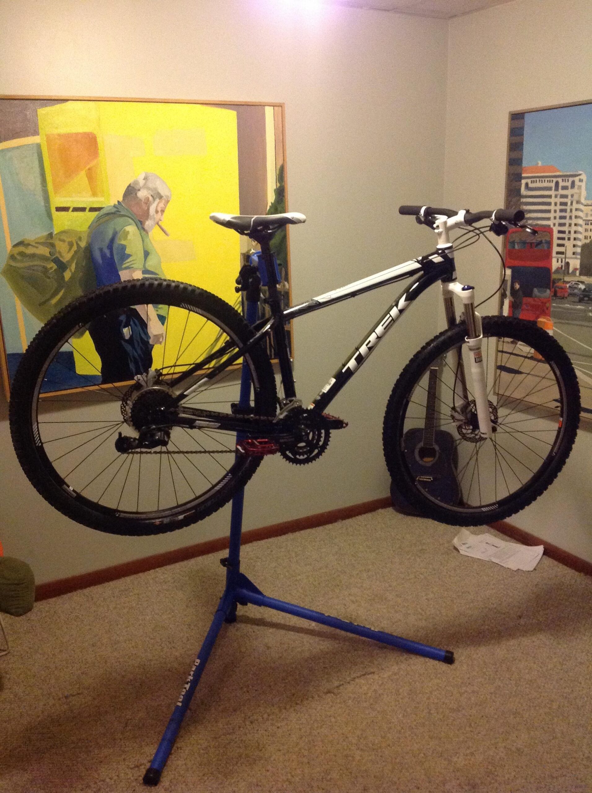 Trek X-Caliber 8: Alt text: A Trek mountain bike is mounted on a blue repair stand in a room with beige carpet. A colorful painting of a man with a cigarette and a yellow background is visible on the wall behind the bike. There is a guitar leaning against the wall and some papers on the floor.