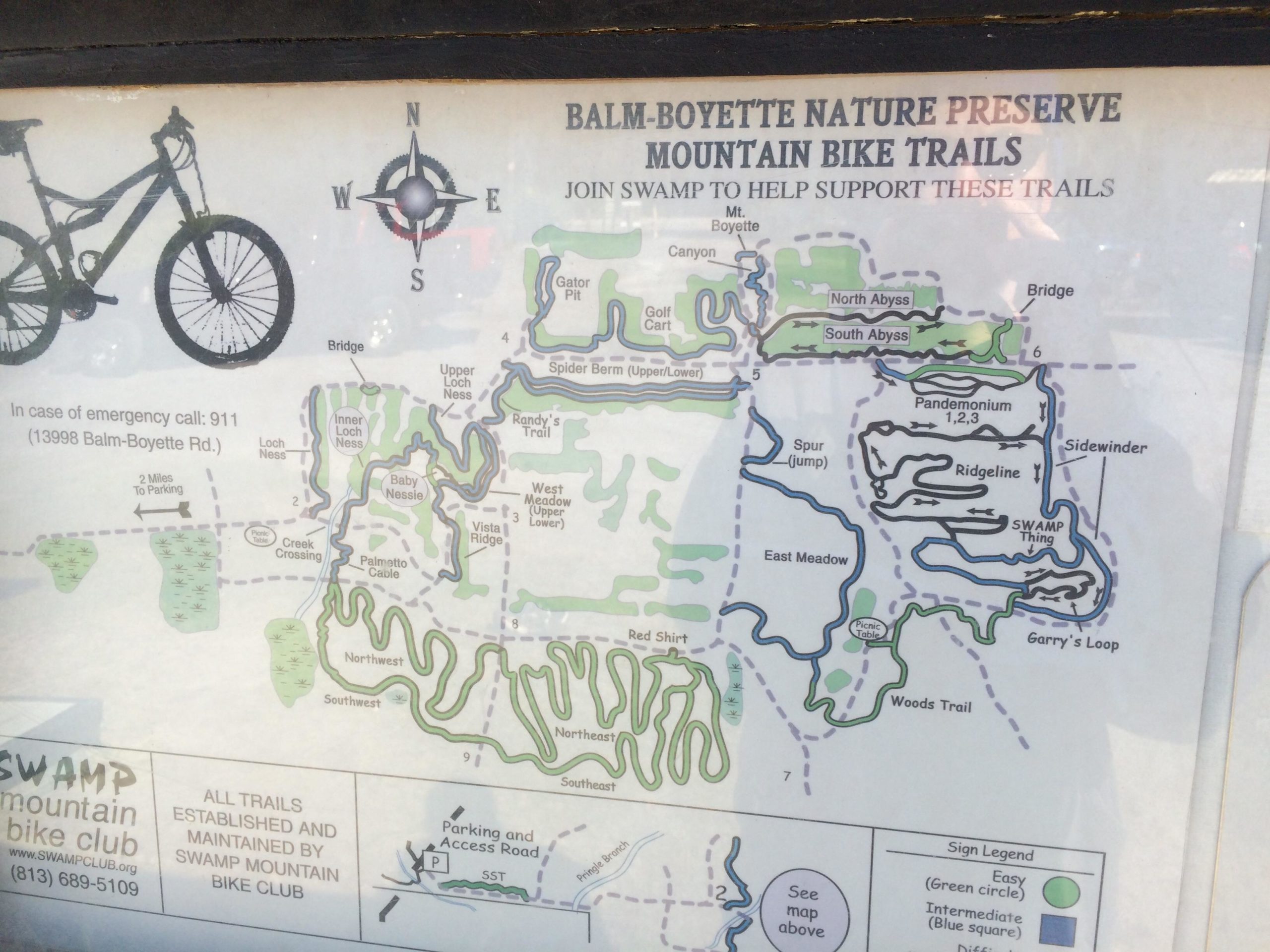 A map of the Balm-Boyette Nature Preserve showcasing the mountain bike trails. The map includes various trails such as "North Abyss," "South Abyss," "Pandemonium," and "Garry's Loop." Key features include a compass rose indicating north, a bike icon, and emergency contact information. The surface level difficulty of each trail is marked using different symbols, with a legend for easy reference. Balm Boyette Scrub Preserve mountain bike trail.