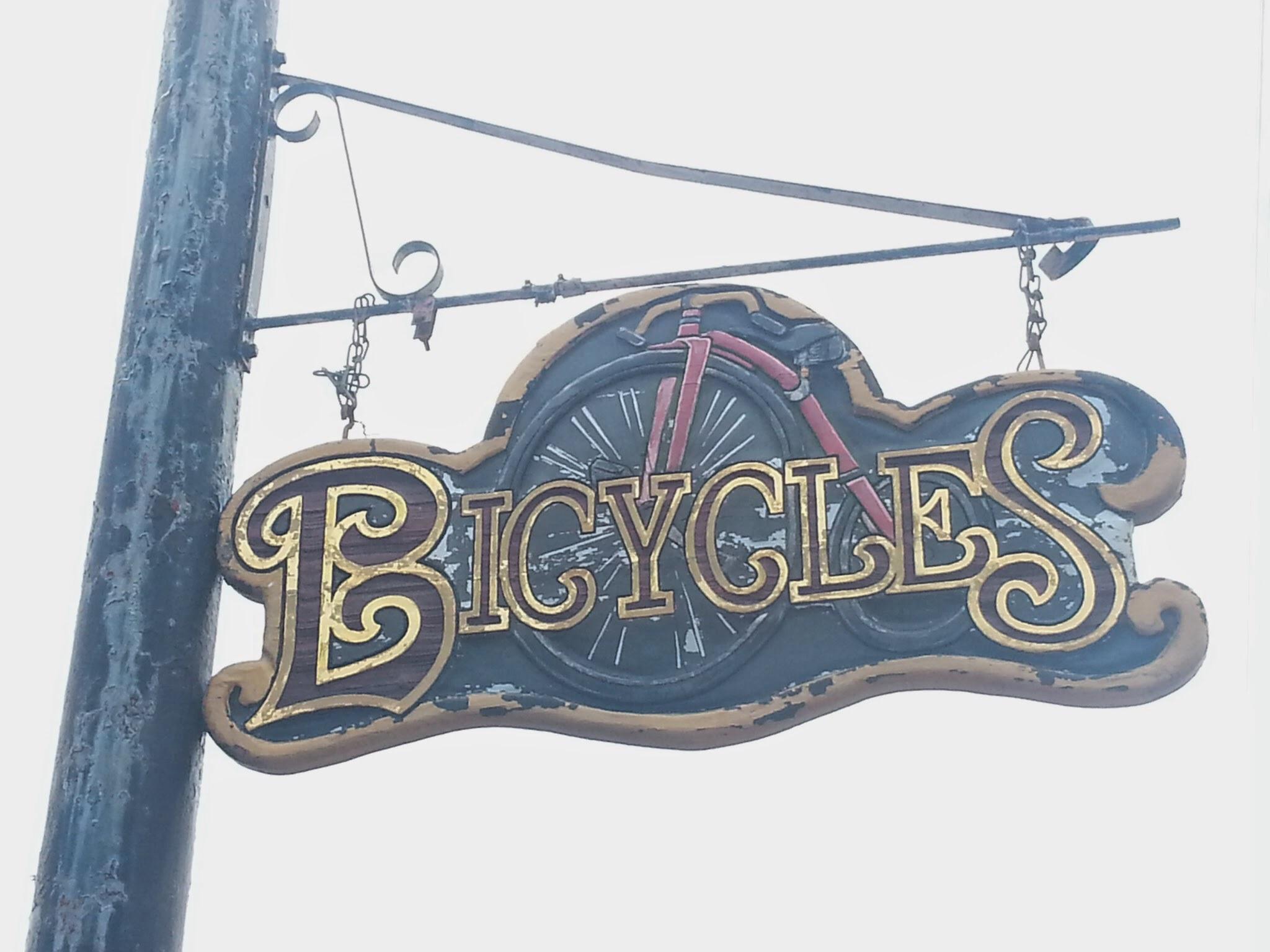 A vintage-style sign hanging from a pole, displaying the word "Bicycles" in decorative lettering with a bicycle wheel integrated into the design. The sign shows signs of wear, adding to its charm.